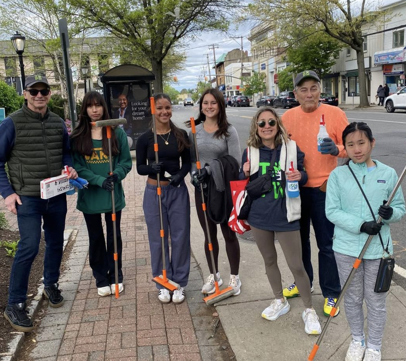 Group of eight diverse volunteers standing on sidewalk with litter pickers and cleaning supplies during a community cleanup day on a city street.