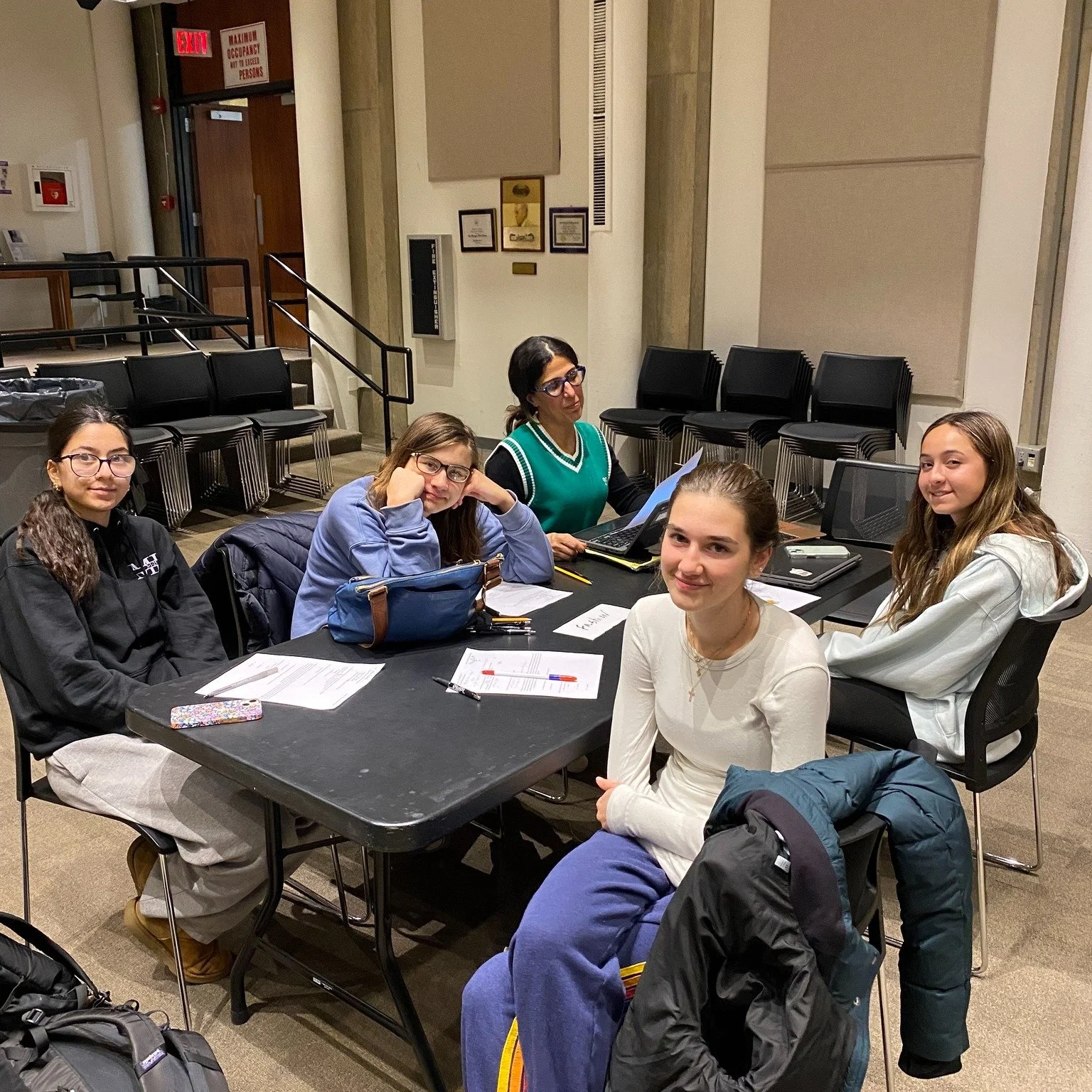Five young women sitting around a black table in a room with beige walls and a stack of black chairs in the background. They appear to be studying or working together, with papers, notebooks, and writing utensils on the table.