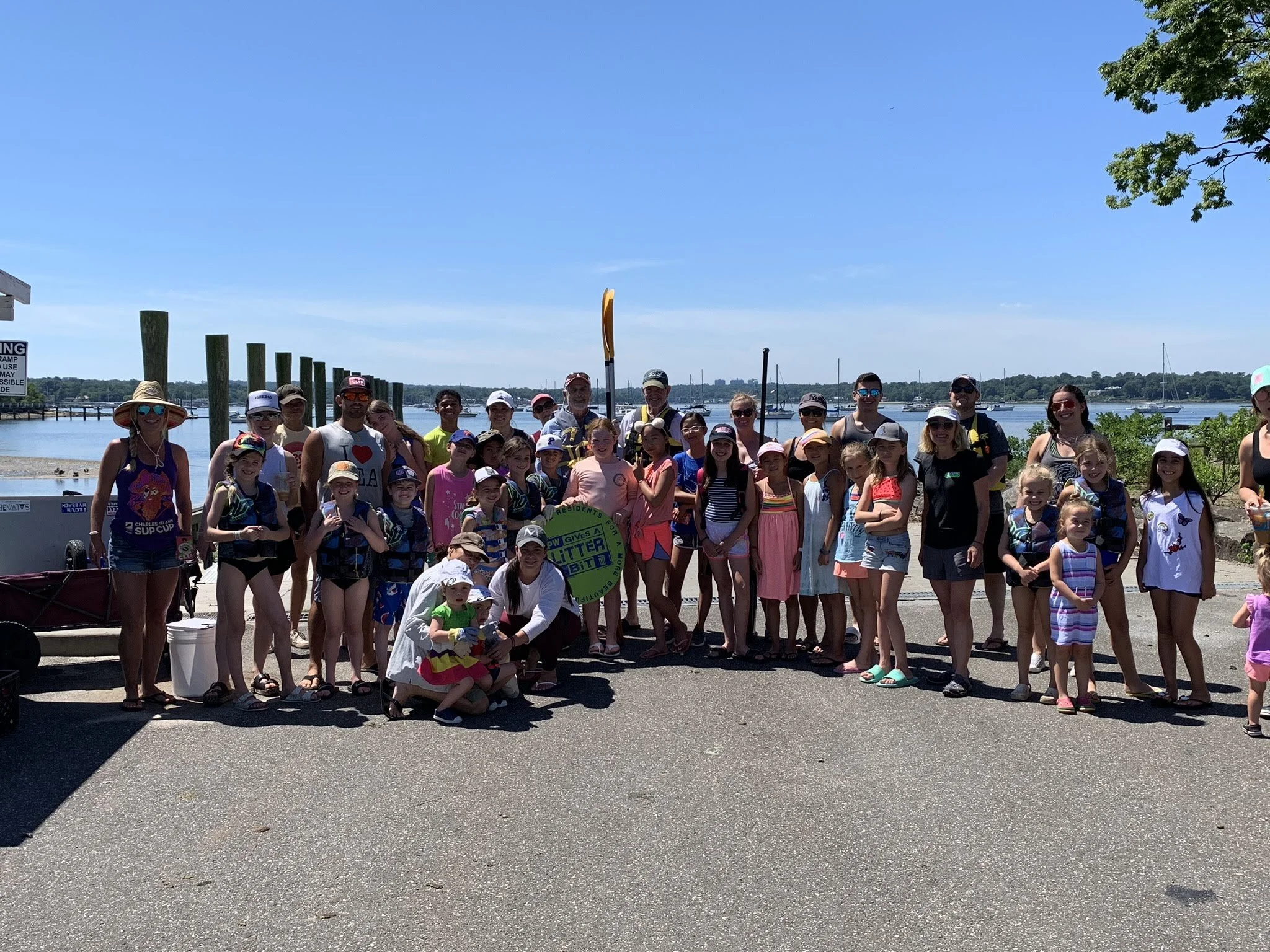 A large group of children and adults standing together outdoors near a waterfront on a sunny day, with boats and water in the background.