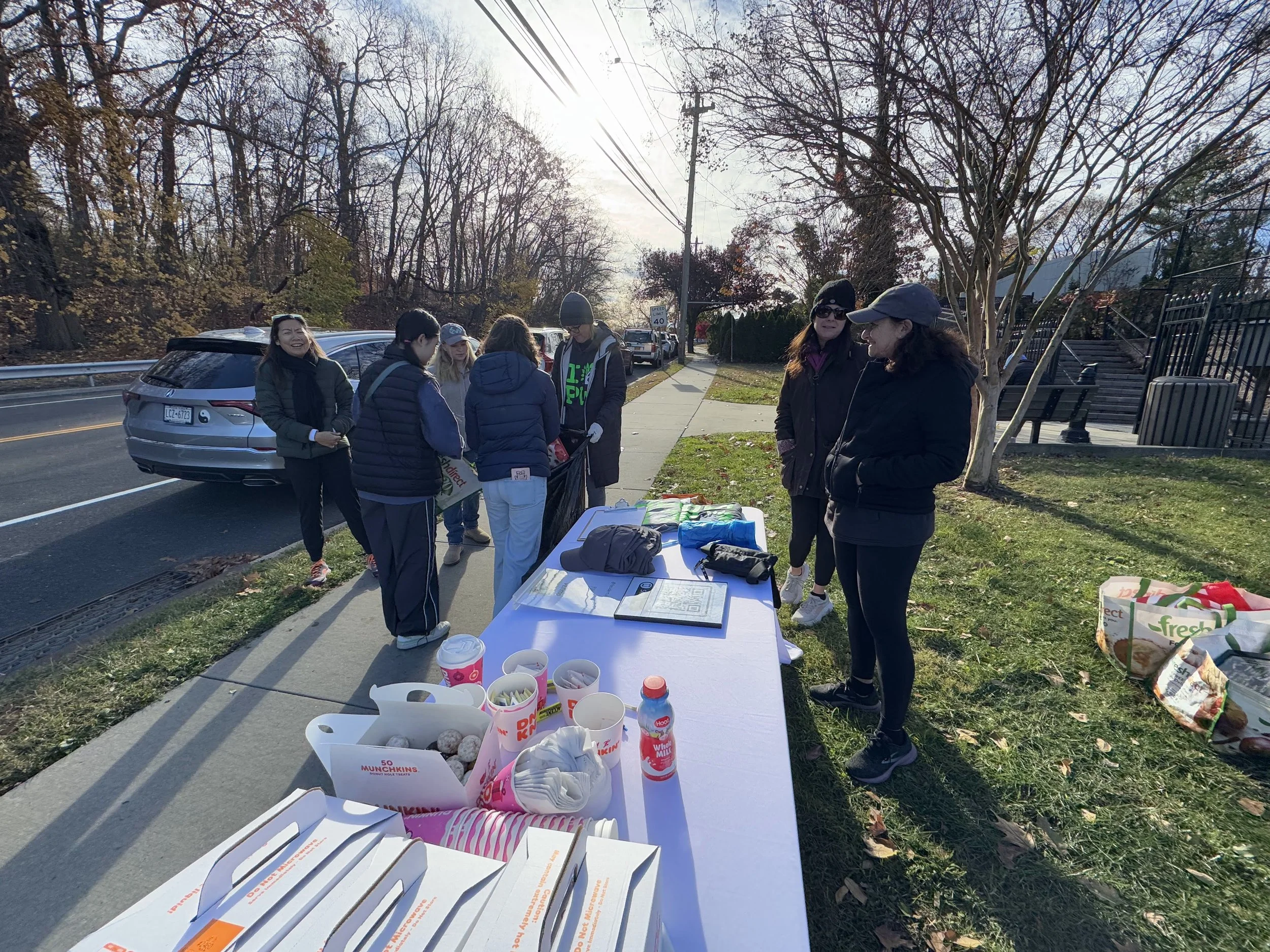 A group of people gathered around a table outdoors on a sidewalk, with some items including baked goods, drinks, and bags. The scene is in a suburban area with trees and a street in the background, on a sunny day.
