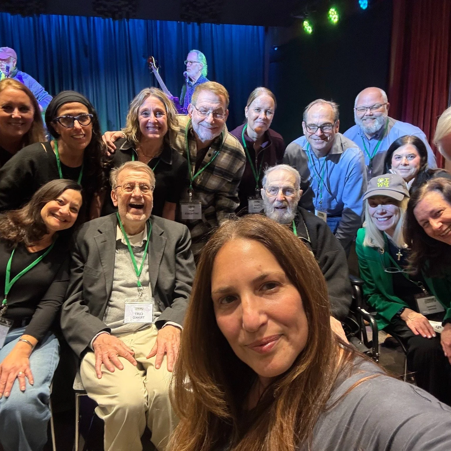 Group of people at an indoor event, posing for a selfie in front of a stage with musical performers, all wearing conference name badges and green lanyards, with a blue curtain backdrop.