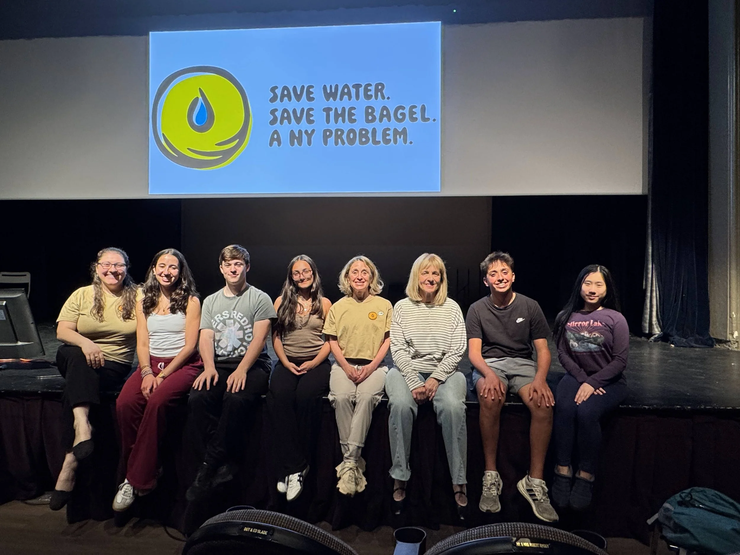 Group of nine people sitting on a stage in front of a large screen displaying an environmental message. The message includes a green circular logo and blue text that says, 'Save water. Save the bagel. A NY problem.'