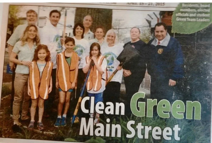 Group of children and adults at a community cleanup event holding trash pickers, with a banner reading 'Clean Green Main Street'.