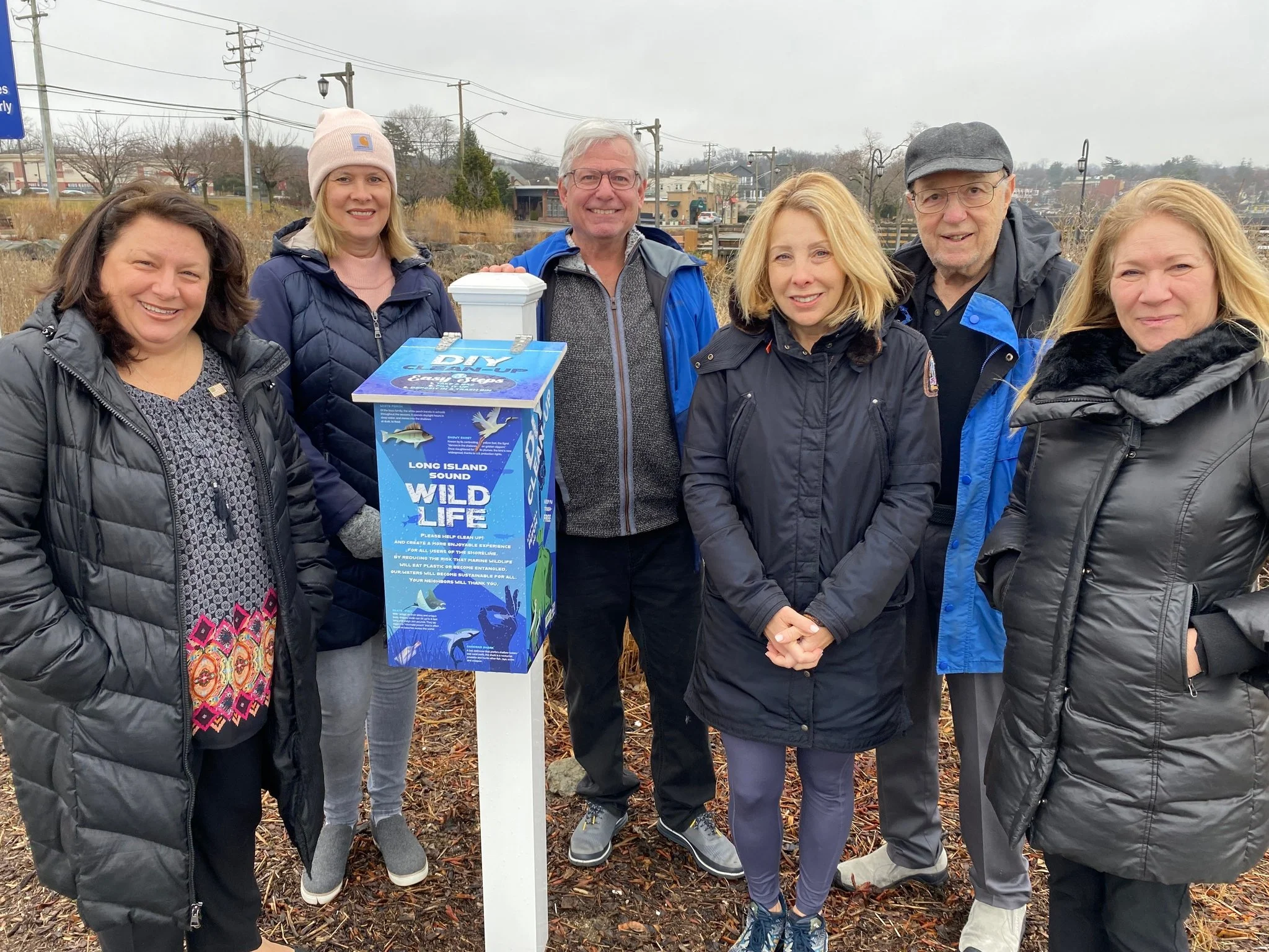 Group of six people standing outdoors next to a blue and white sign that says 'Long Island Sound Wild Life' and has a bird graphic, with a background of a dock and trees. All are wearing winter jackets.