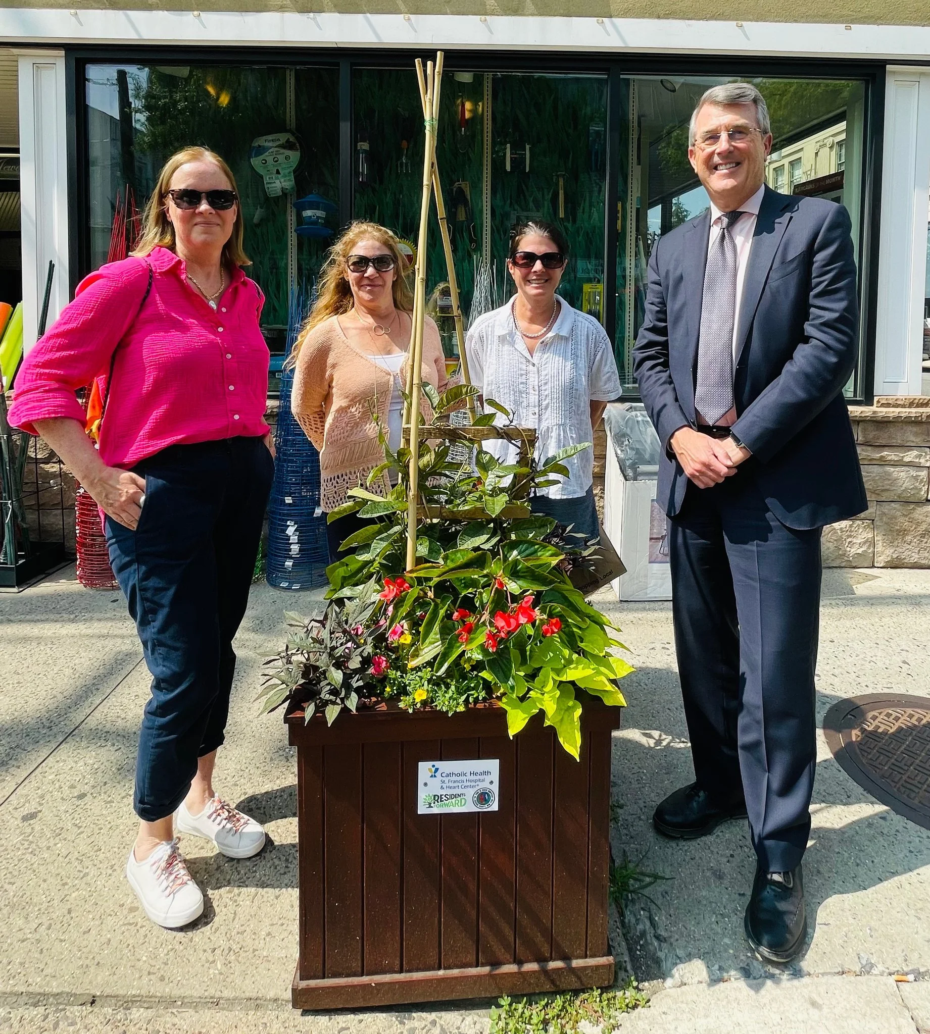 Four people standing behind a large flower arrangement outside a building. One woman is wearing a pink shirt and sunglasses, another woman in beige, a third woman in white, and a man in a suit. The flower arrangement is in a wooden container with a sign that reads 'Catholic Health'.