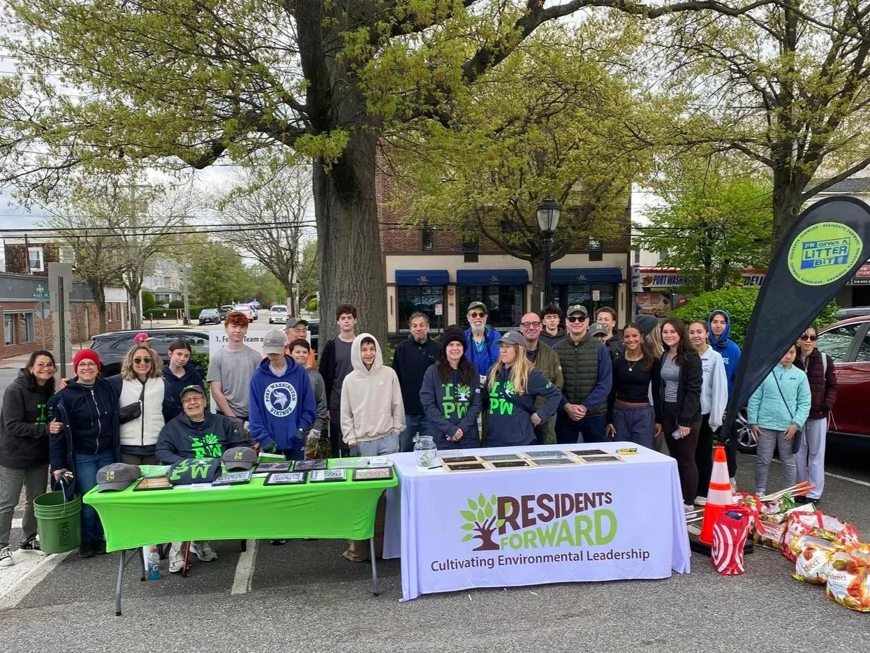 Group of people gathered outdoors around tables with environmental organization banners, trees, and parked cars in the background.