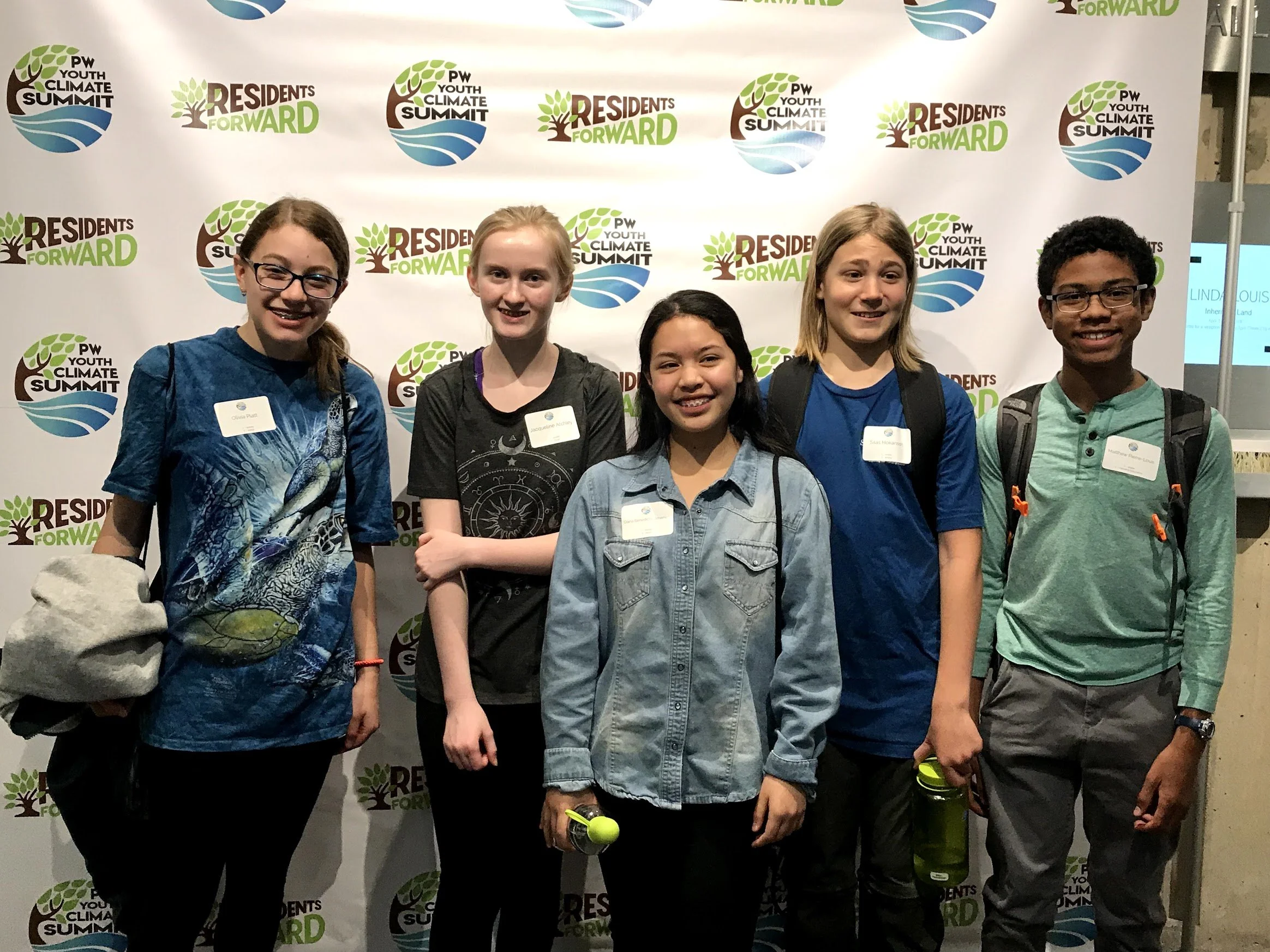 Group of five young people standing in front of a backdrop with logos for the PW Youth Climate Summit and Residents Forward, smiling at the camera.