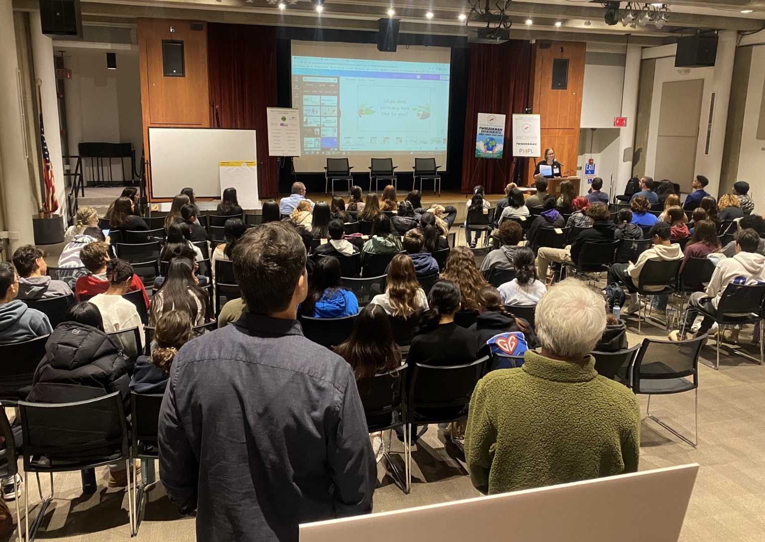 A large group of diverse people attending a presentation or conference in an auditorium, with a woman speaking at a podium on stage, a large screen displaying a PowerPoint slide, and banners on the side of the stage.