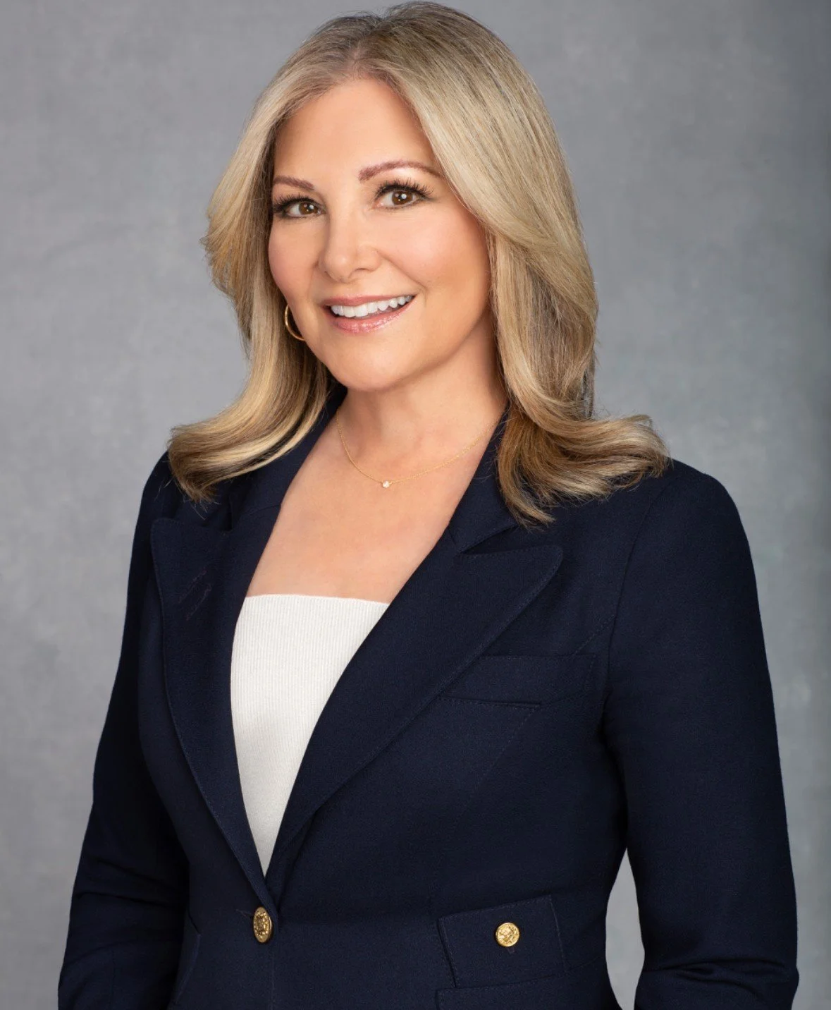 Professional headshot of a woman with blonde hair wearing a navy blazer over a light top, smiling at the camera against a gray background.