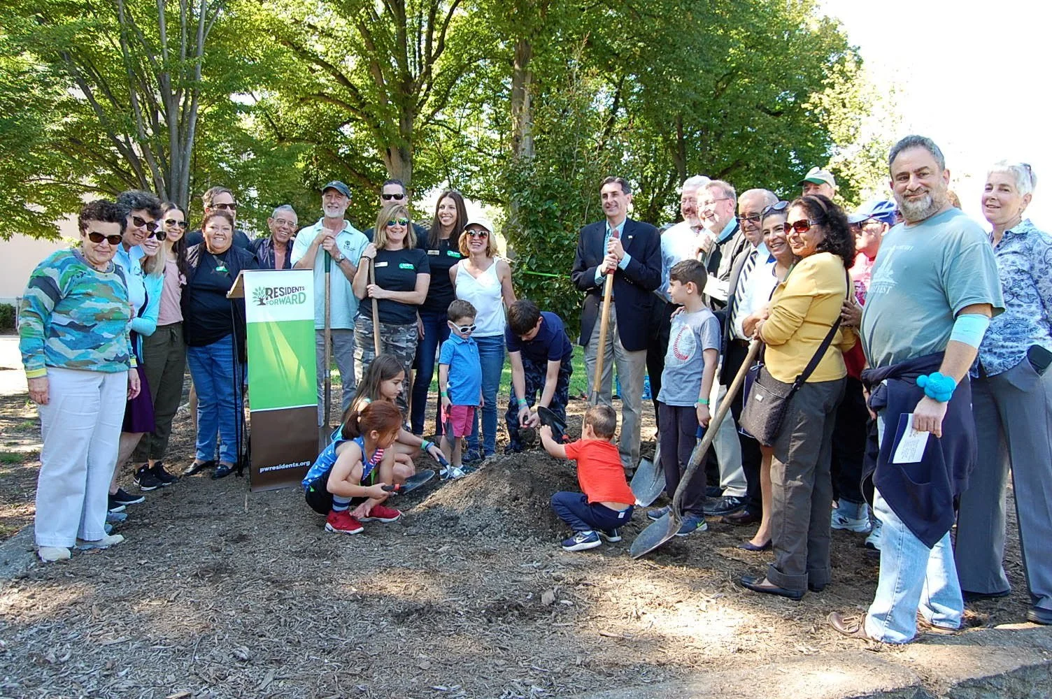 Group of people participating in a tree planting event outdoors, with children and adults holding shovels and gathered around a small mound of soil.