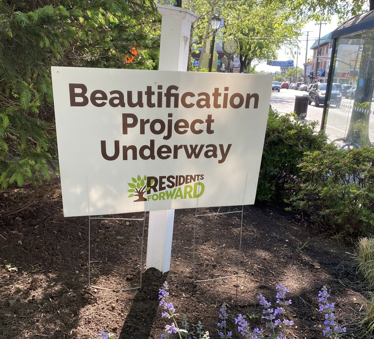 A sign that reads "Beautification Project Underway" with the logo of "Residents Forward" at a landscaped public area with purple flowers, trees, and a busy street in the background.