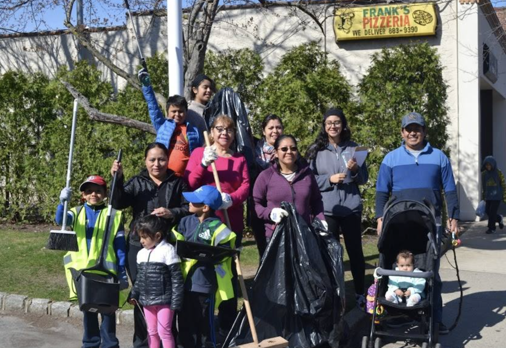 Group of people, including children and adults, participating in a neighborhood clean-up event. They are outdoors in front of a white wall with a sign for a pizzeria. The group is holding trash bags, brooms, and trash pickers, and wearing gloves and 