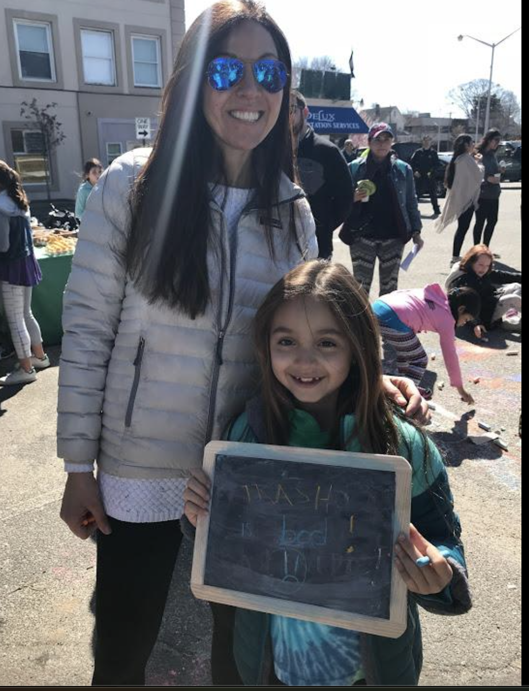 Smiling woman and girl holding a small chalkboard with writing, at an outdoor community event on a sunny day, with other people and activities in the background.