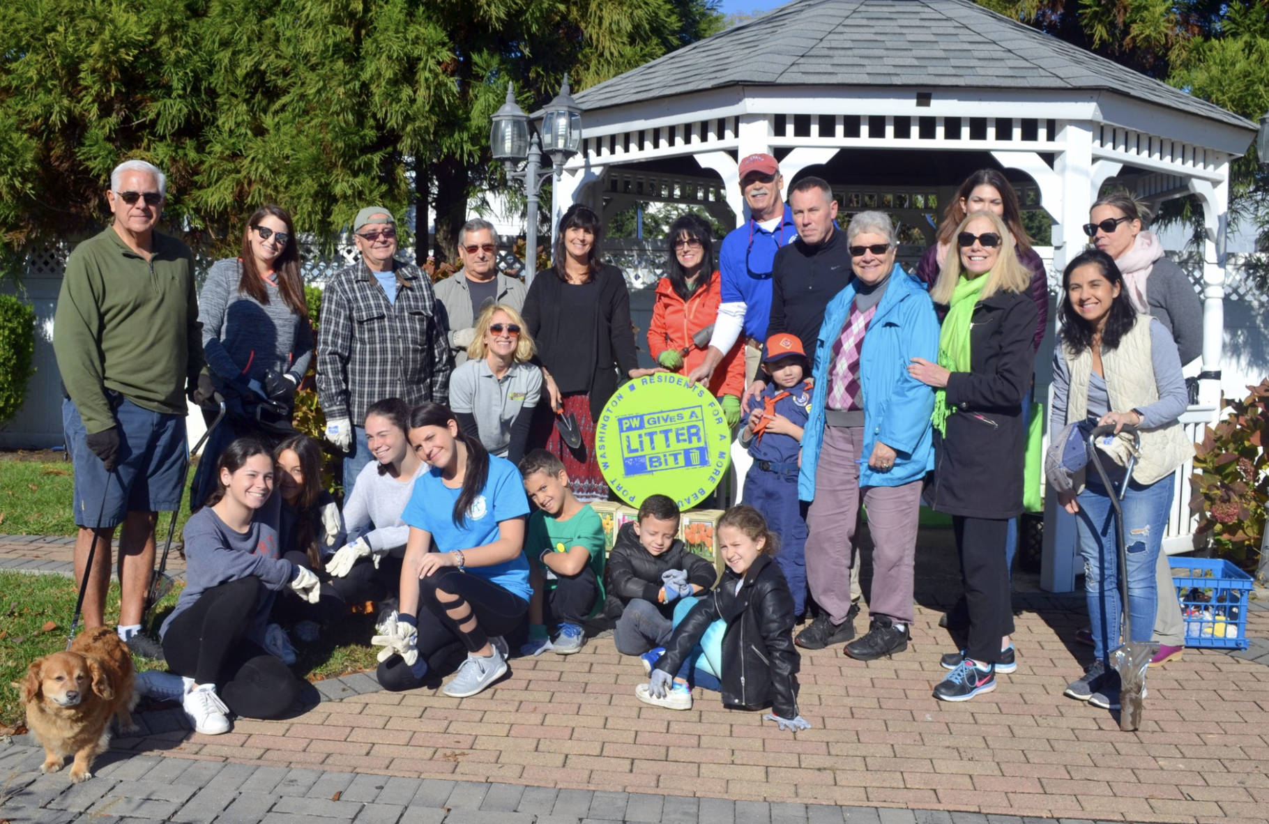 Group of people participating in a community cleanup event, holding a sign that reads 'PW GIVES A LITTER BIT', in front of a white gazebo with trees in the background.
