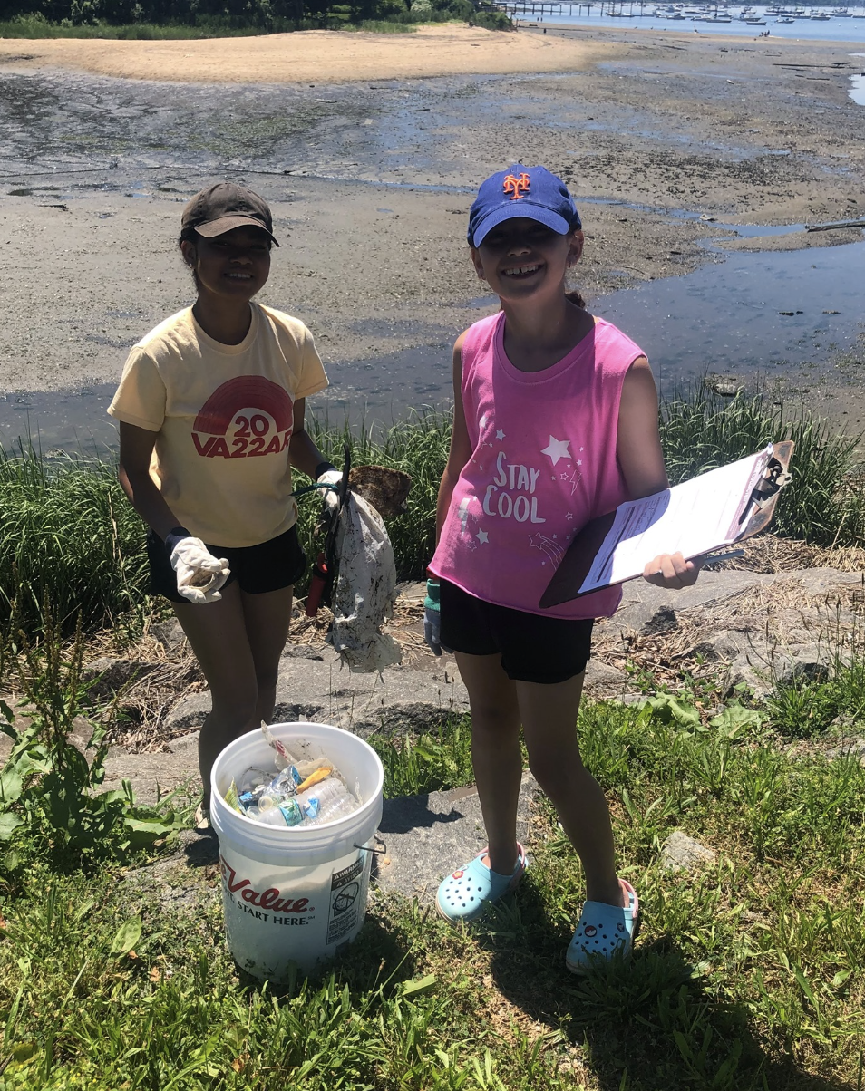 Two young girls collecting trash by the water's edge. One girl is holding a trash bag and wearing a cap and a yellow T-shirt. The other girl is holding a clipboard, wearing a blue cap, pink sleeveless shirt, and Crocs. There is a bucket of collected 