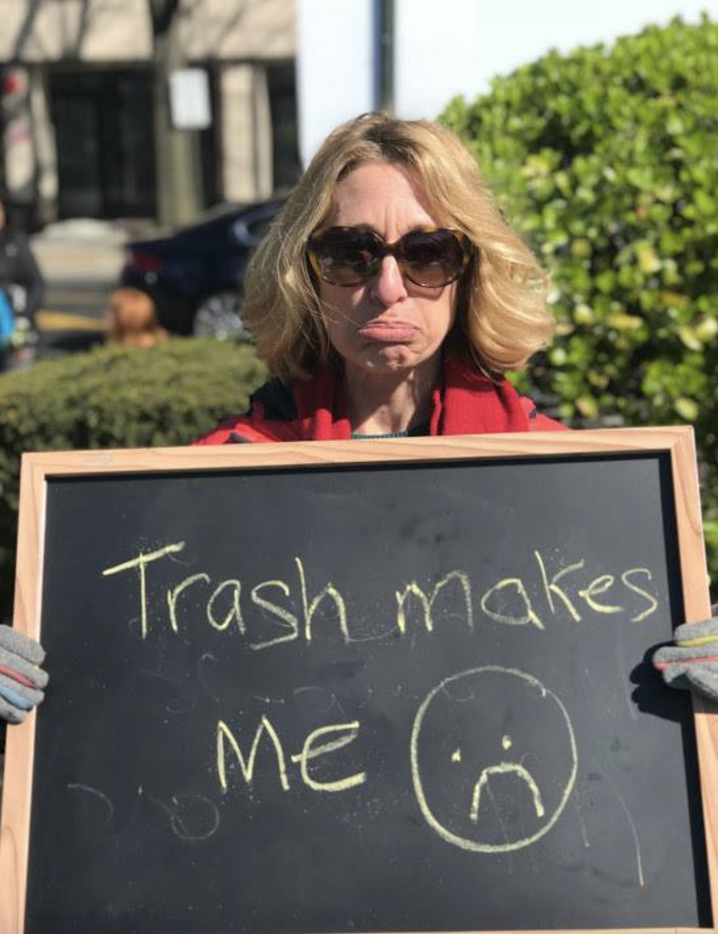 A woman with blonde hair, wearing sunglasses and a red jacket, holding a blackboard with the words "Trash makes ME" and a sad face drawn on it.