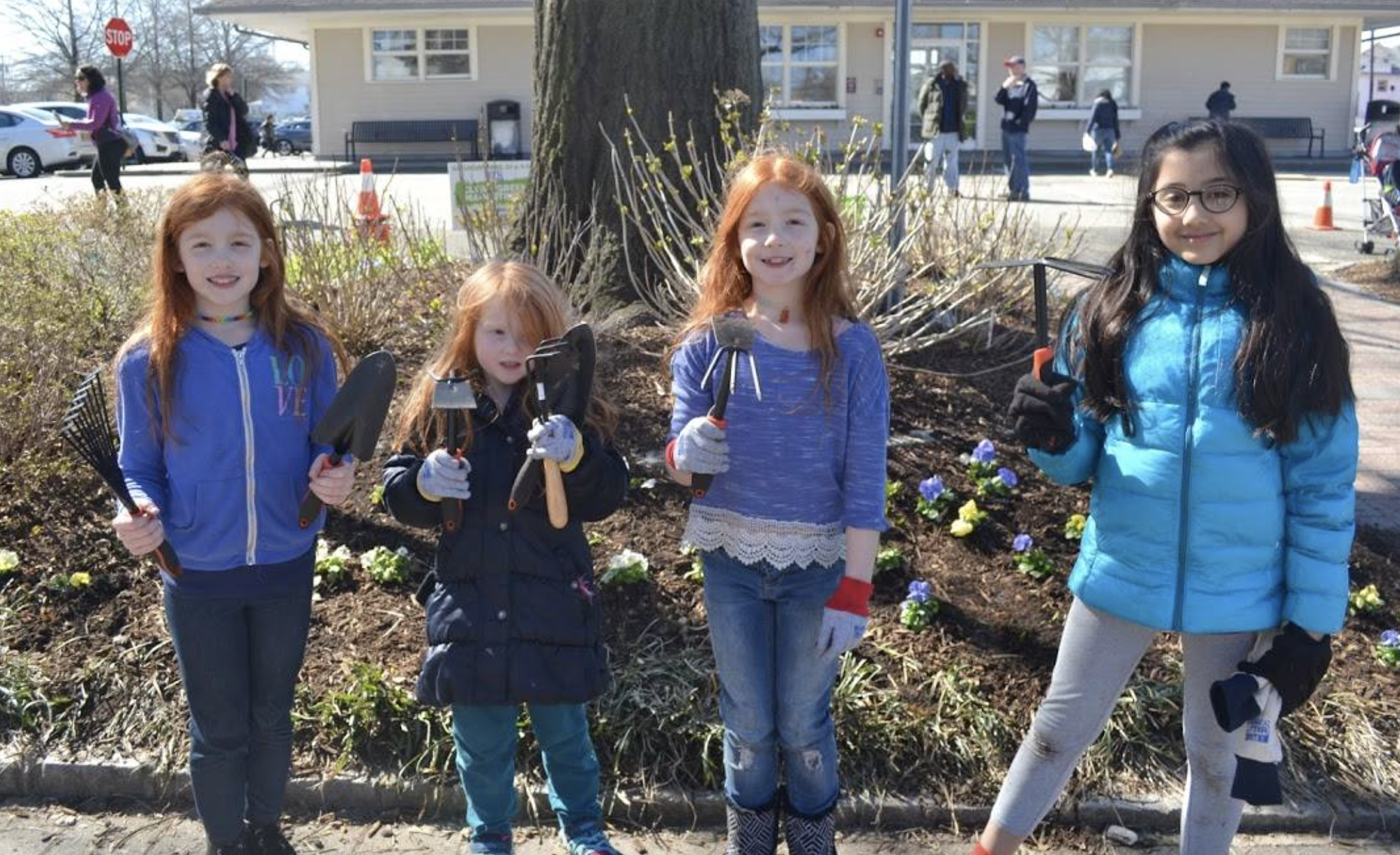Four girls stand in front of a flower bed, holding gardening tools and wearing gloves, smiling outdoors on a sunny day.