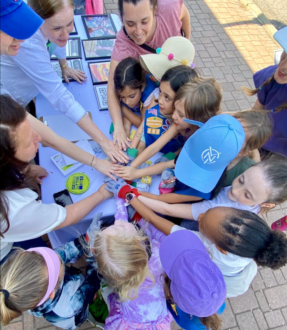 A group of children and adults gathered around a table, placing their hands together in the center, participating in a fun outdoor activity. The table has various items including tablets and a yellow sign that says "LITTER." The children are wearing 