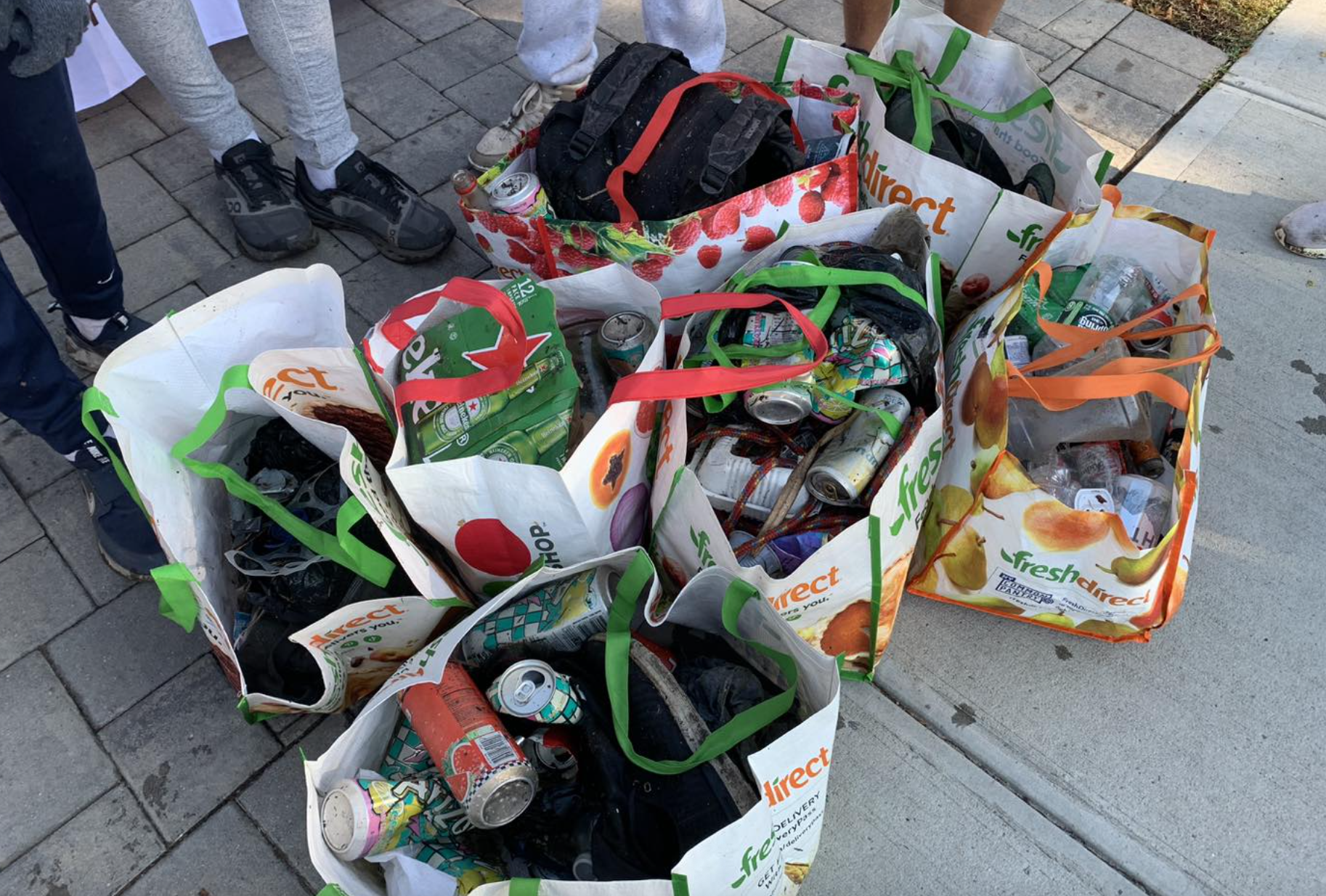 Recycling bags filled with cans, bottles, and waste, placed on a sidewalk, with people standing around.