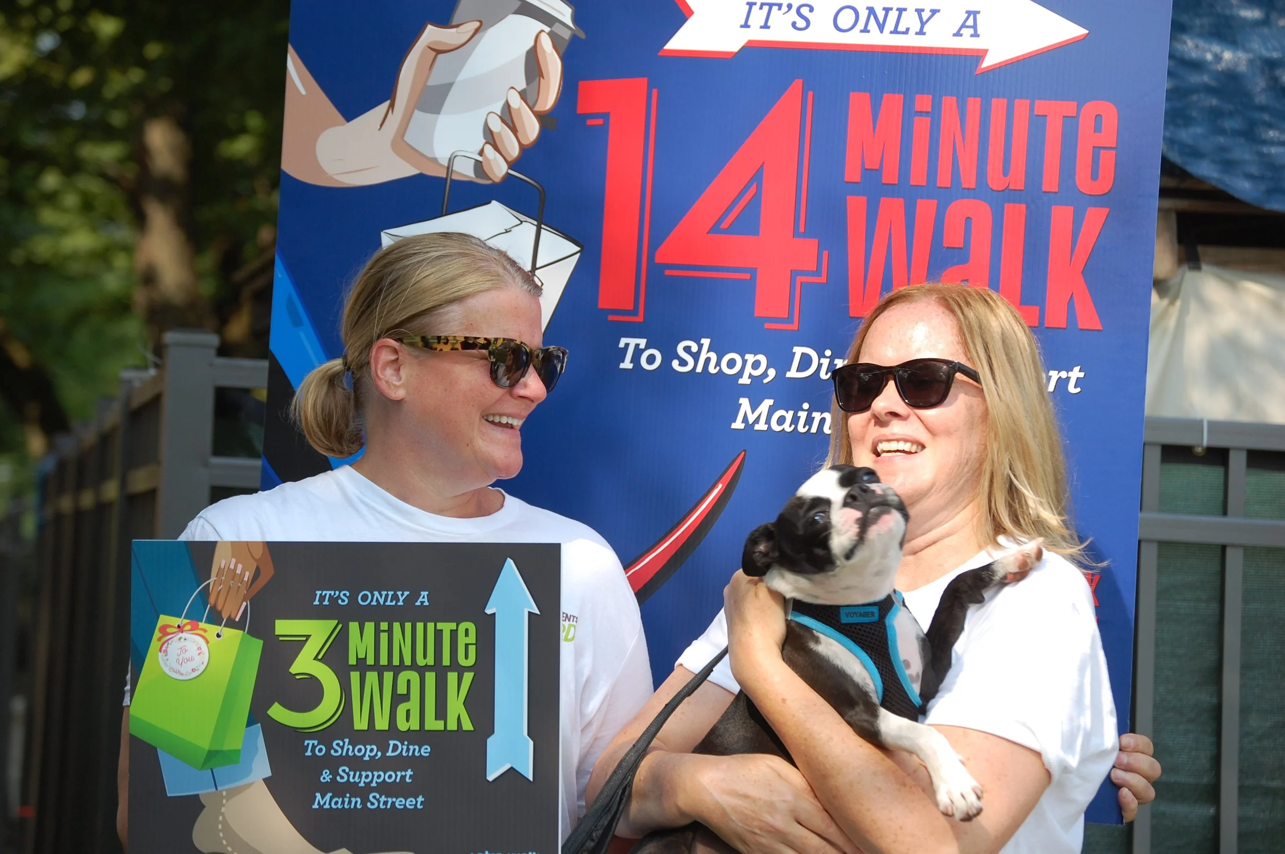 Two women smiling at each other, one holding a small Boston Terrier puppy, standing in front of colorful signs promoting community walks for shopping and dining support.