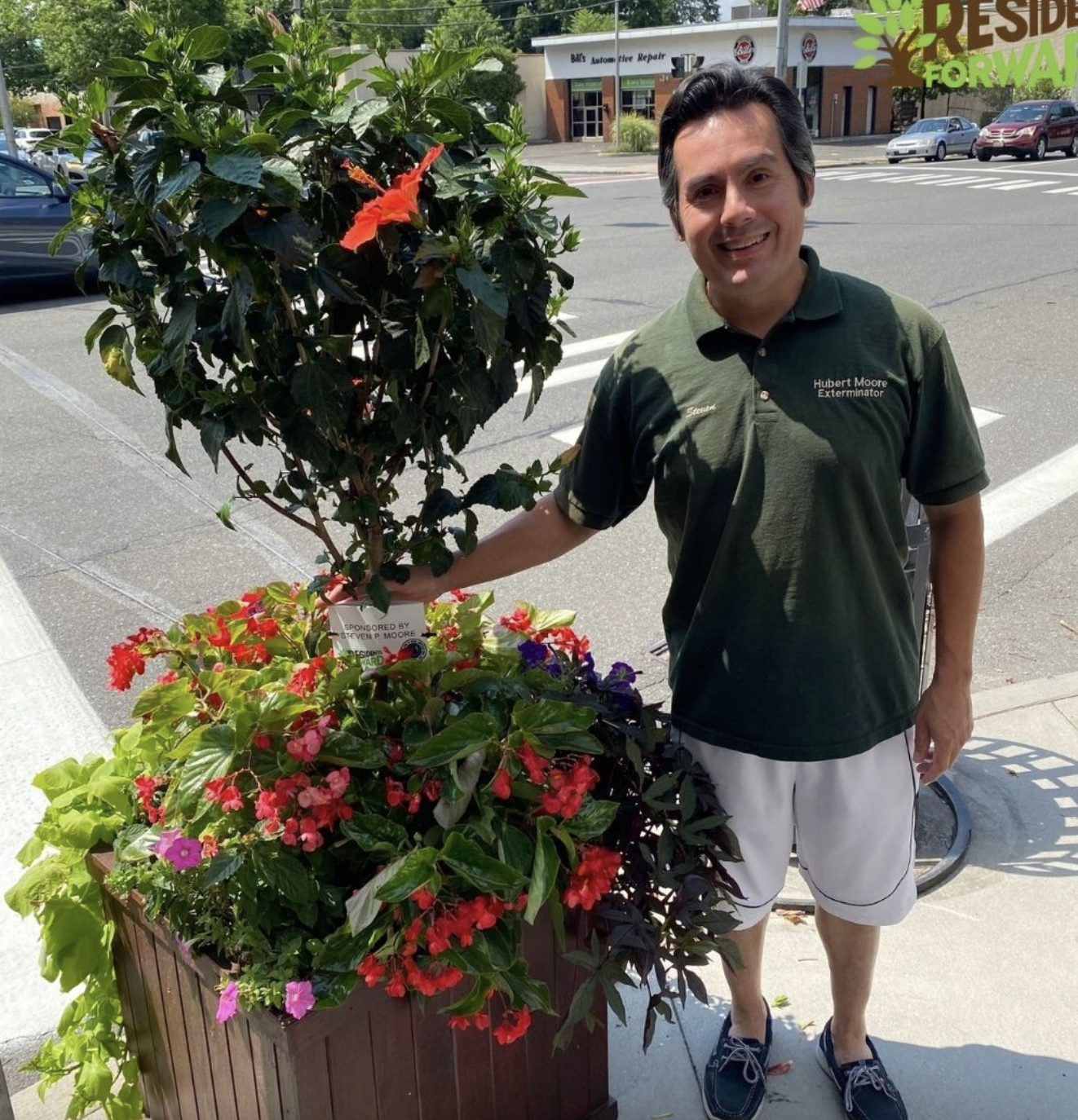 A man standing next to a large flower pot containing various colorful flowers, on a sidewalk in an urban area. He is smiling and wearing a green polo shirt and shorts.