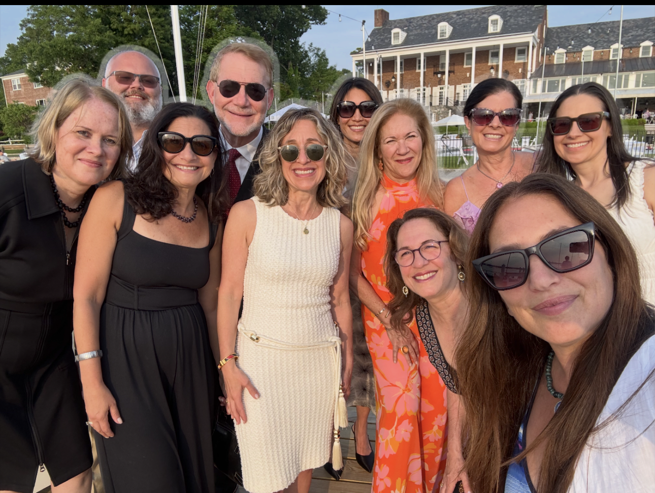 Ten people posing together outdoors at a social gathering, smiling and wearing sunglasses, with a large brick building and trees in the background.