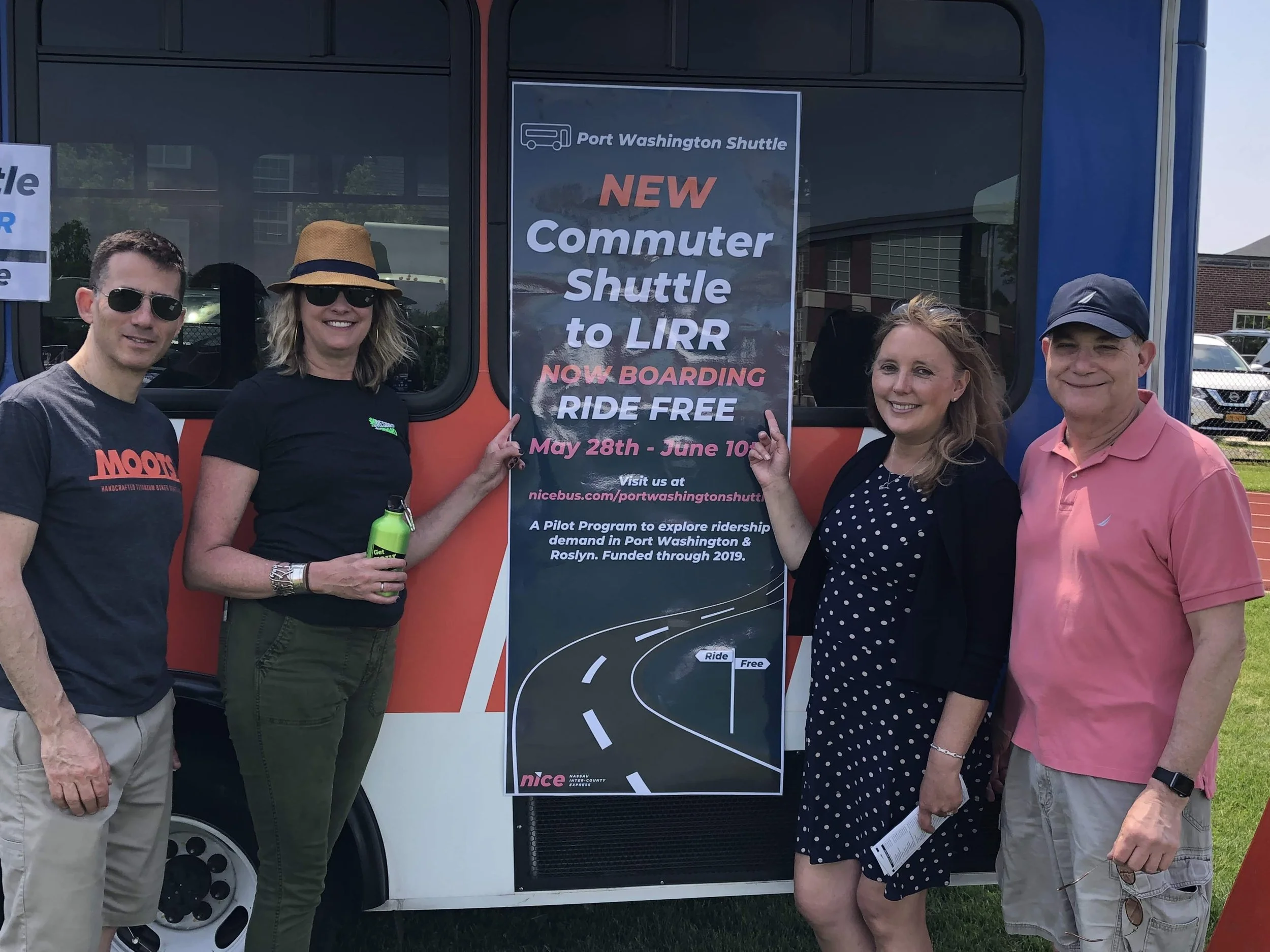 Four people standing outdoors next to a bus with a sign promoting a new commuter shuttle to LIRR. The sign mentions the schedule from May 28th to June 10th and that riding is free. The group includes two women and two men, all smiling and casually dressed, with some wearing sunglasses.