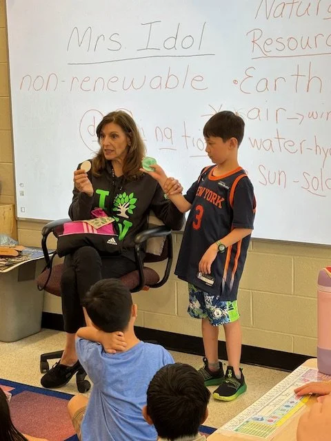 A classroom scene with a female teacher sitting in a chair and a young male student standing next to her, holding a globe. Other students are seated on the floor, facing the teacher. Behind them is a whiteboard with notes about Mrs. Idol, non-renewable resources, and Earth.