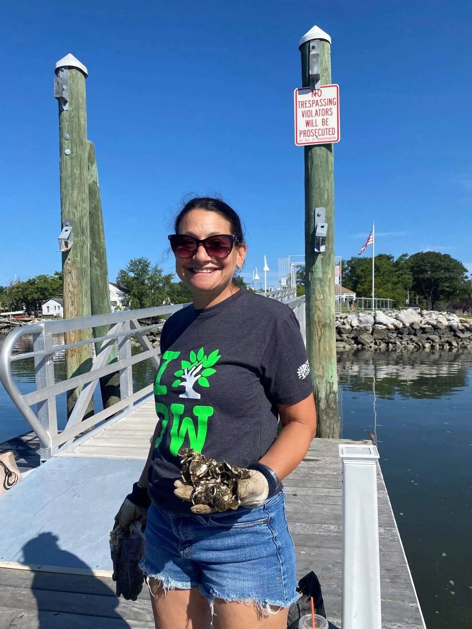 A woman with sunglasses, a black T-shirt with a green and white tree logo, and denim shorts standing on a dock by the water, holding a cluster of oysters with a smile.