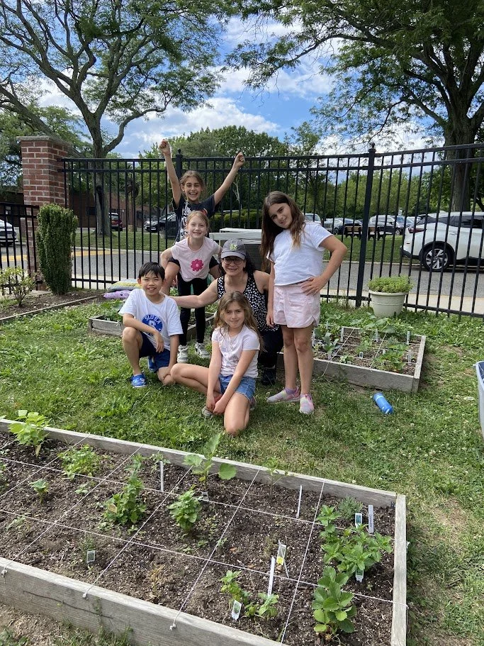 Six children, four girls and two boys, are smiling and posing next to a vegetable garden with plants on a sunny day. They are outdoors, with trees, a black fence, parked cars, and a partly cloudy sky in the background.