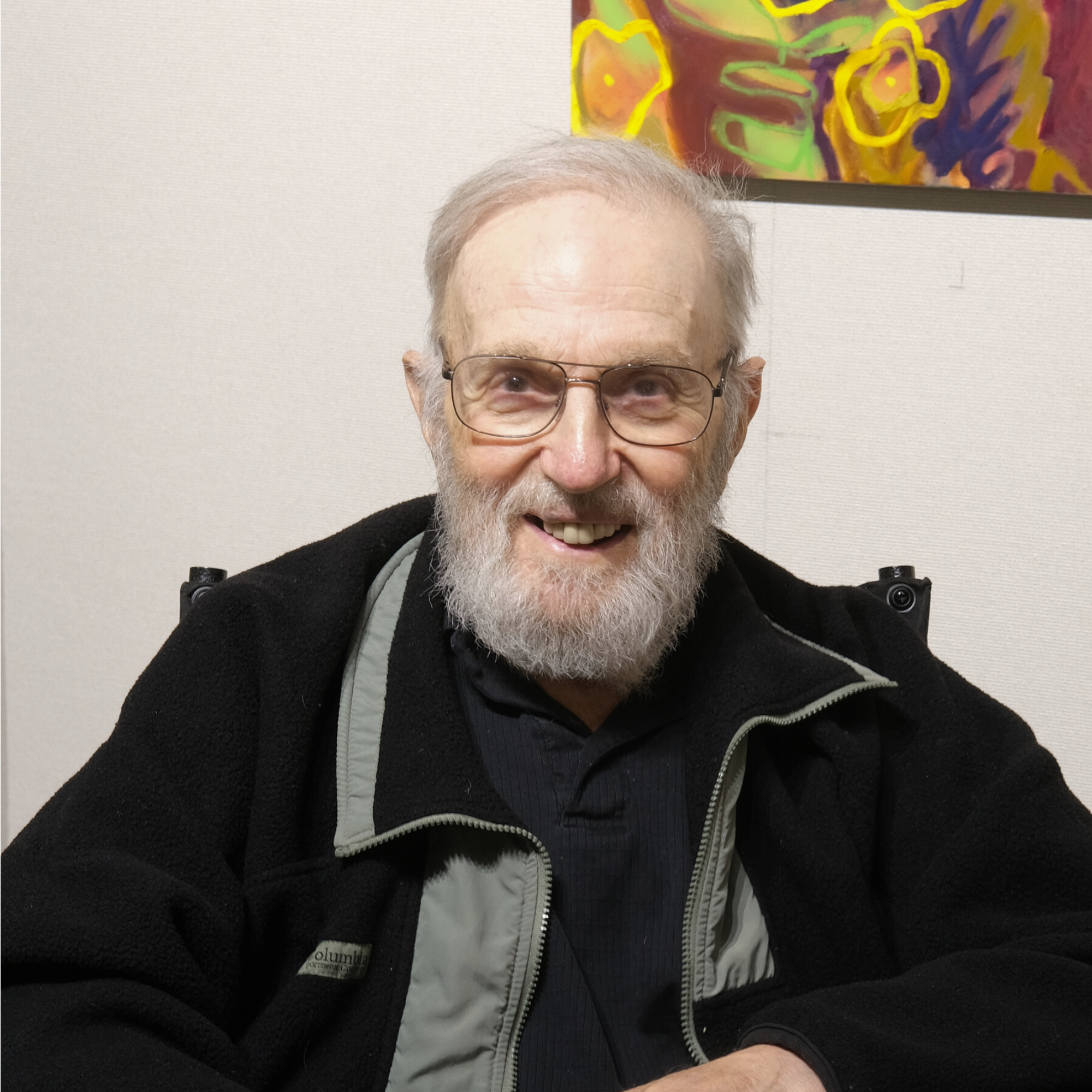 An elderly man with glasses and a beard smiling while sitting in front of a white wall with colorful abstract art.