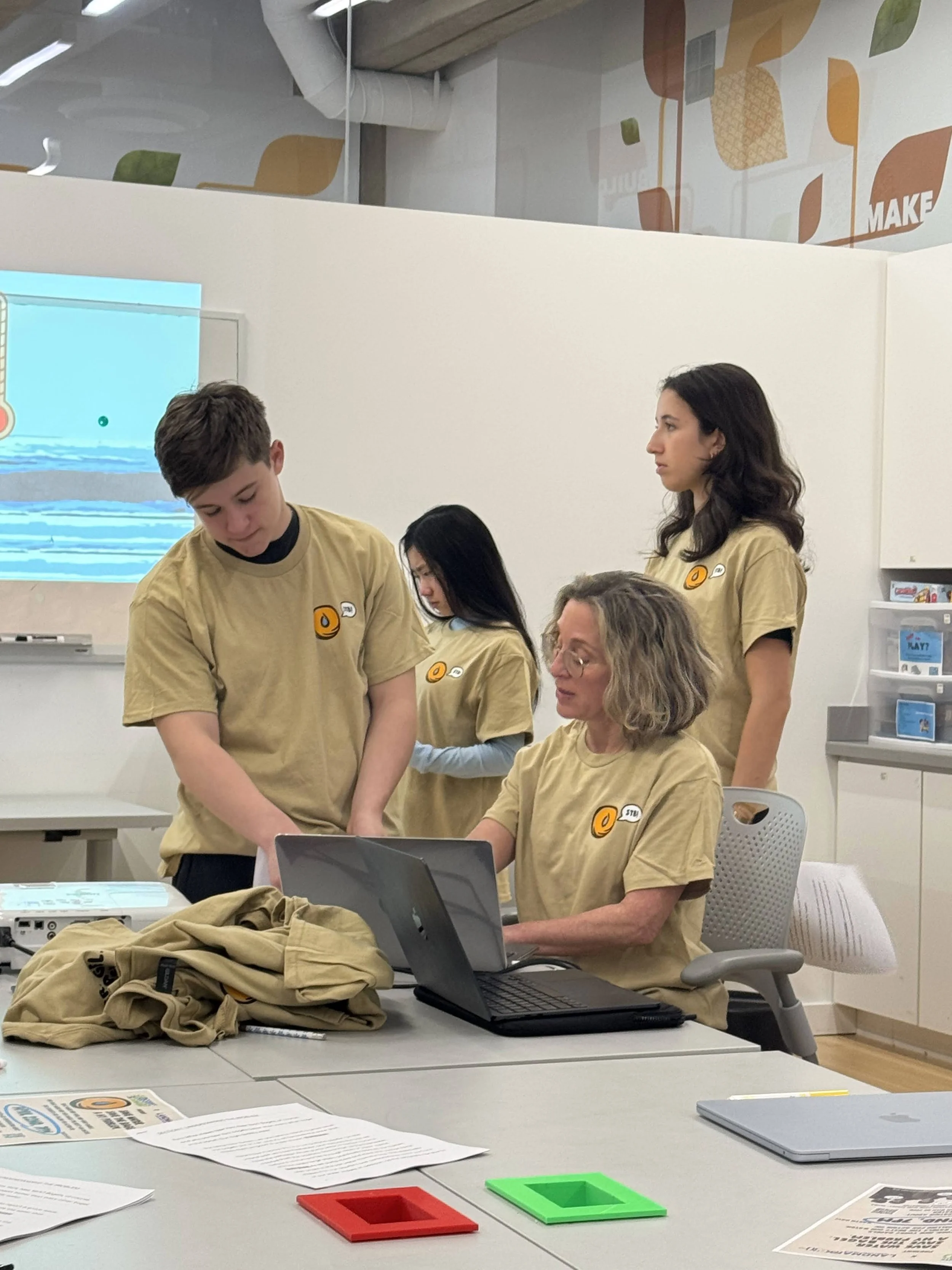 A group of four people in beige shirts with logo patches in a classroom or workshop, gathered around a laptop, with one person seated and three standing, engaged in a discussion or instructions.