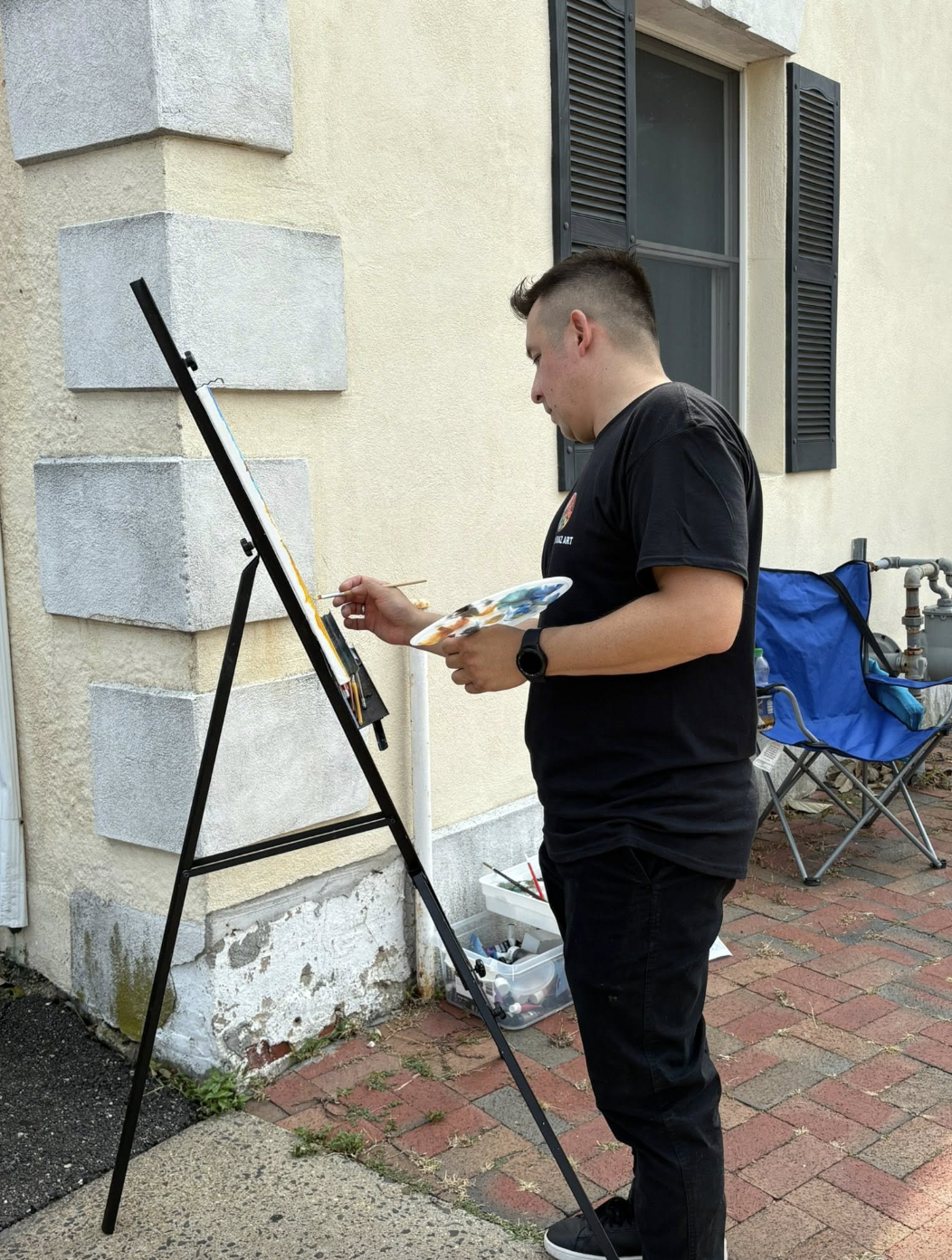 A young man painting on an outdoor easel beside a house wall.