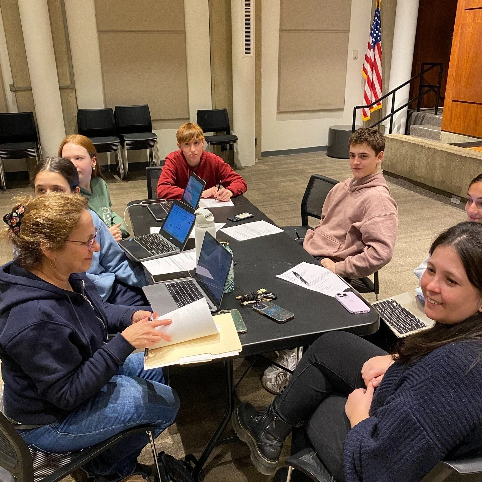 Group of people sitting around a table with laptops, notebooks, and papers in a conference room.