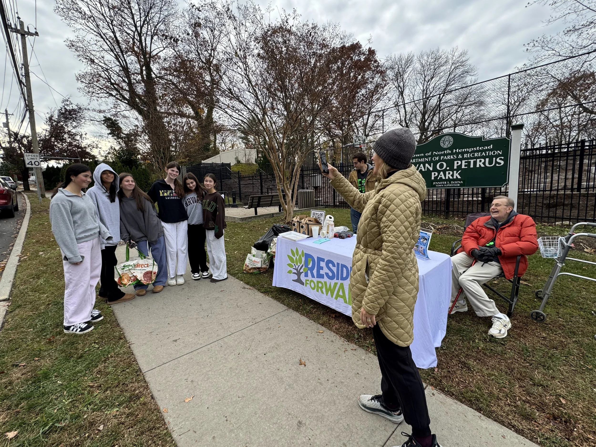 Group of young women receiving food donations from volunteers at a community event in North Hempstead, New York. The event is part of a community outreach program at a park with a table displaying the Residence Forward banner. The background features