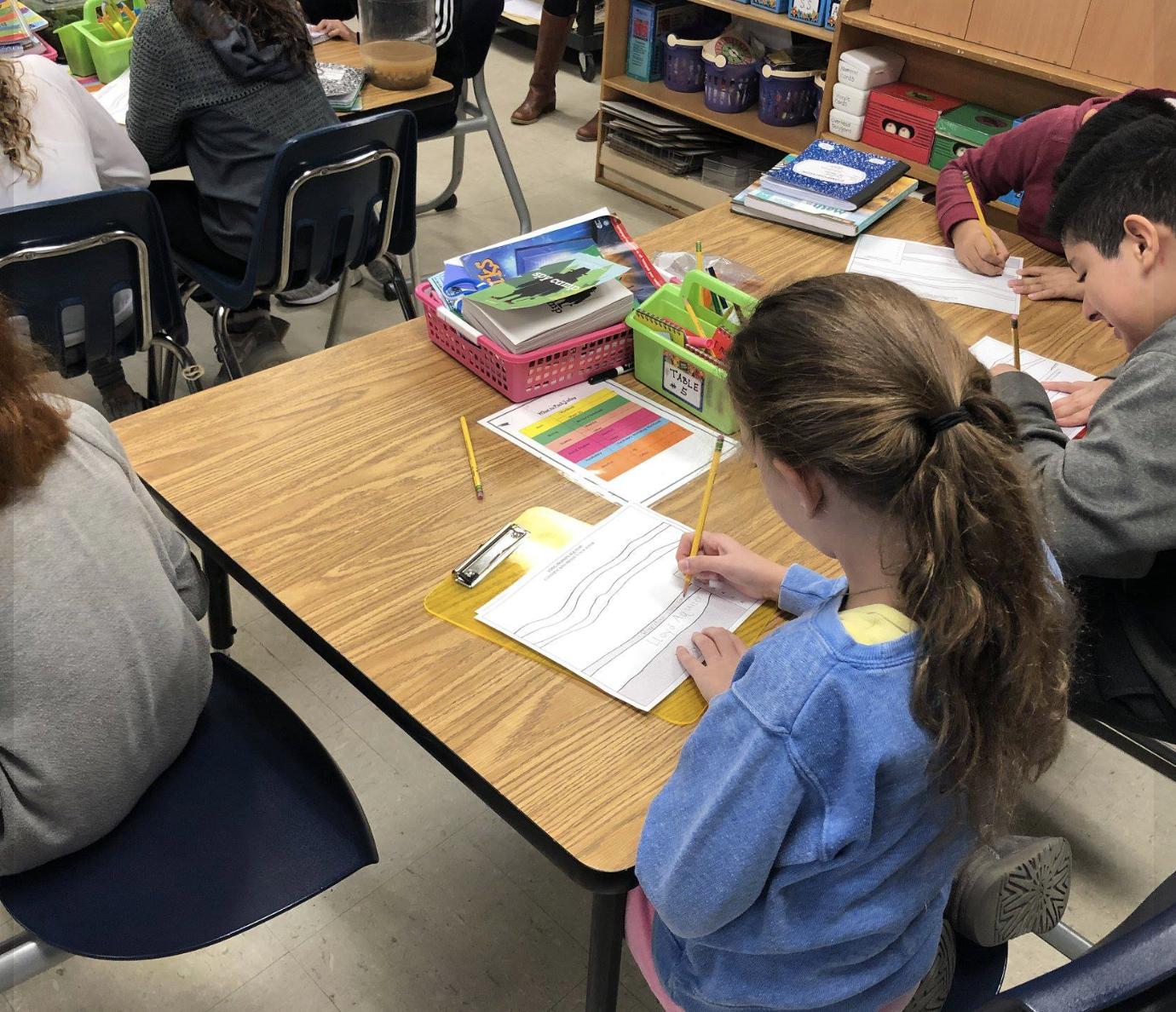 A young girl with brown curly hair wearing a blue sweatshirt, sitting at a wooden classroom table, coloring in a workbook with a yellow pencil. Other students are sitting nearby, working on assignments. The table has various school supplies like colo