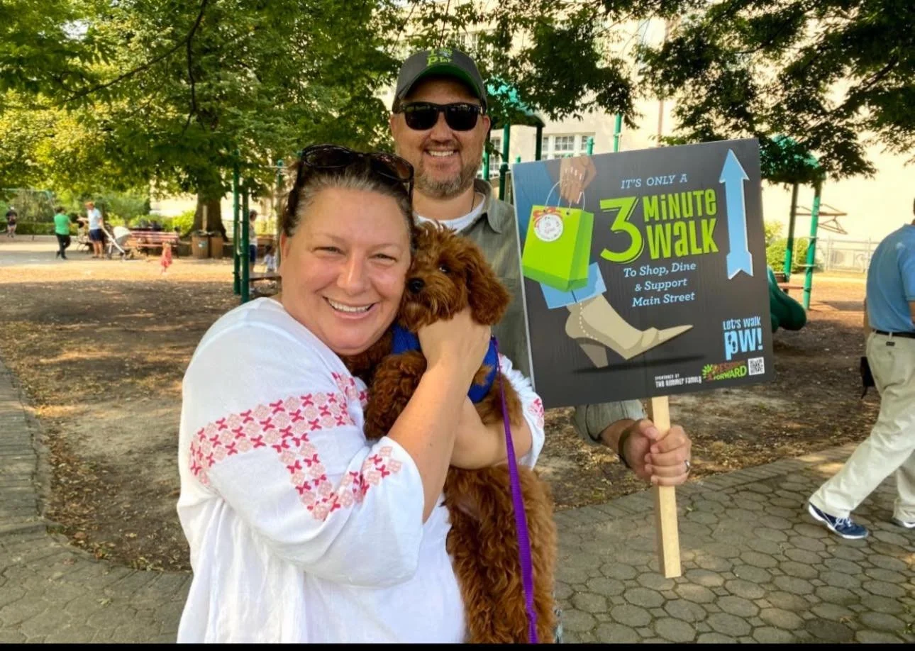 A woman smiling and holding a small brown dog, standing next to a man who is holding a sign that reads 'It's only a 3-minute walk to shop, dine, and support Main Street' during a community walk event in a park with trees and other people in the backg