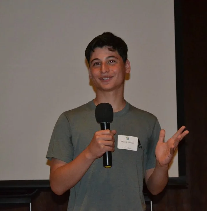 A young boy holding a microphone and speaking, wearing a green t-shirt and a name tag that says Max Goldman, standing in front of a white screen.