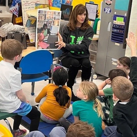 A teacher reading a book to a group of young students in a classroom. The teacher is sitting on a chair with children seated on the floor listening attentively.