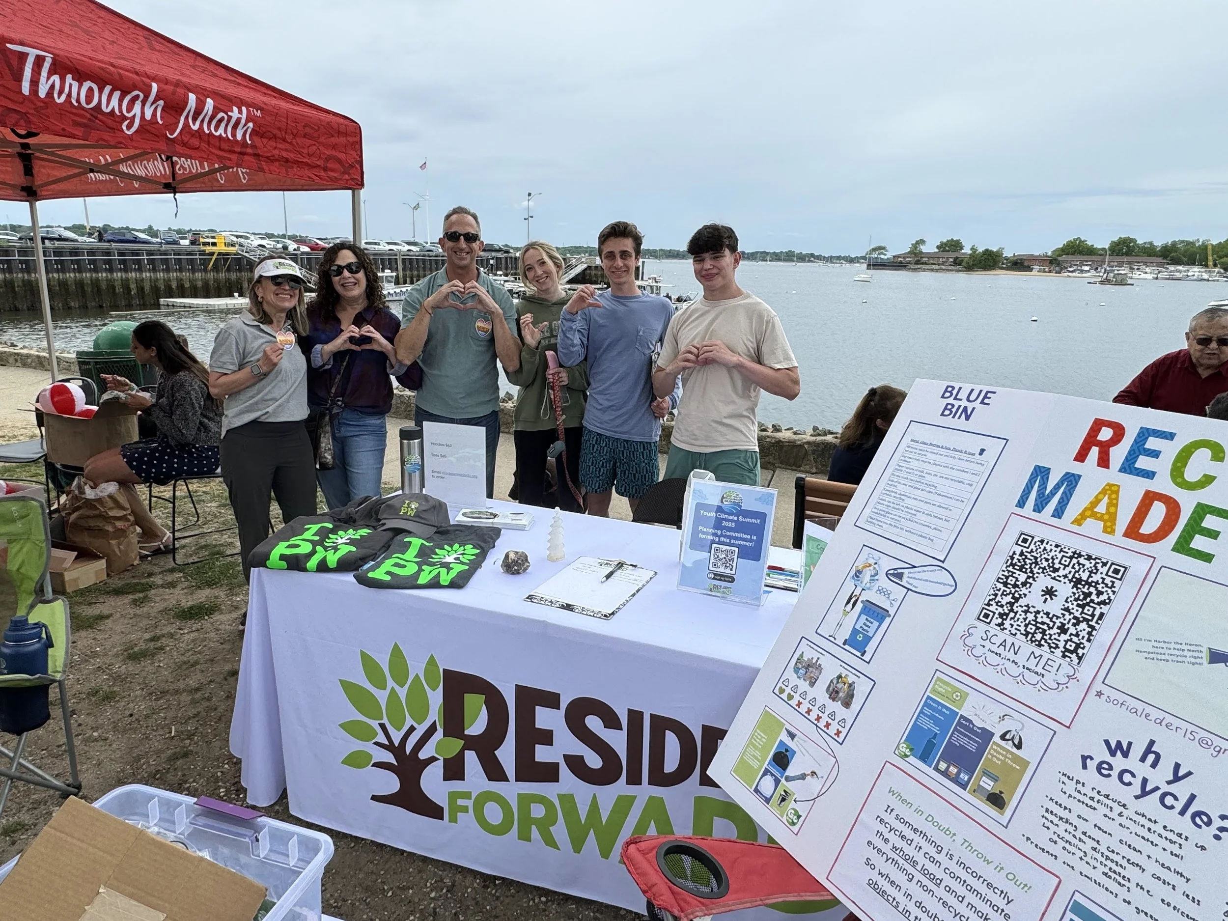 Group of six people standing behind a table at an outdoor event by the water, with some making heart and peace signs with their hands, and a large informational poster about recycling in the foreground.
