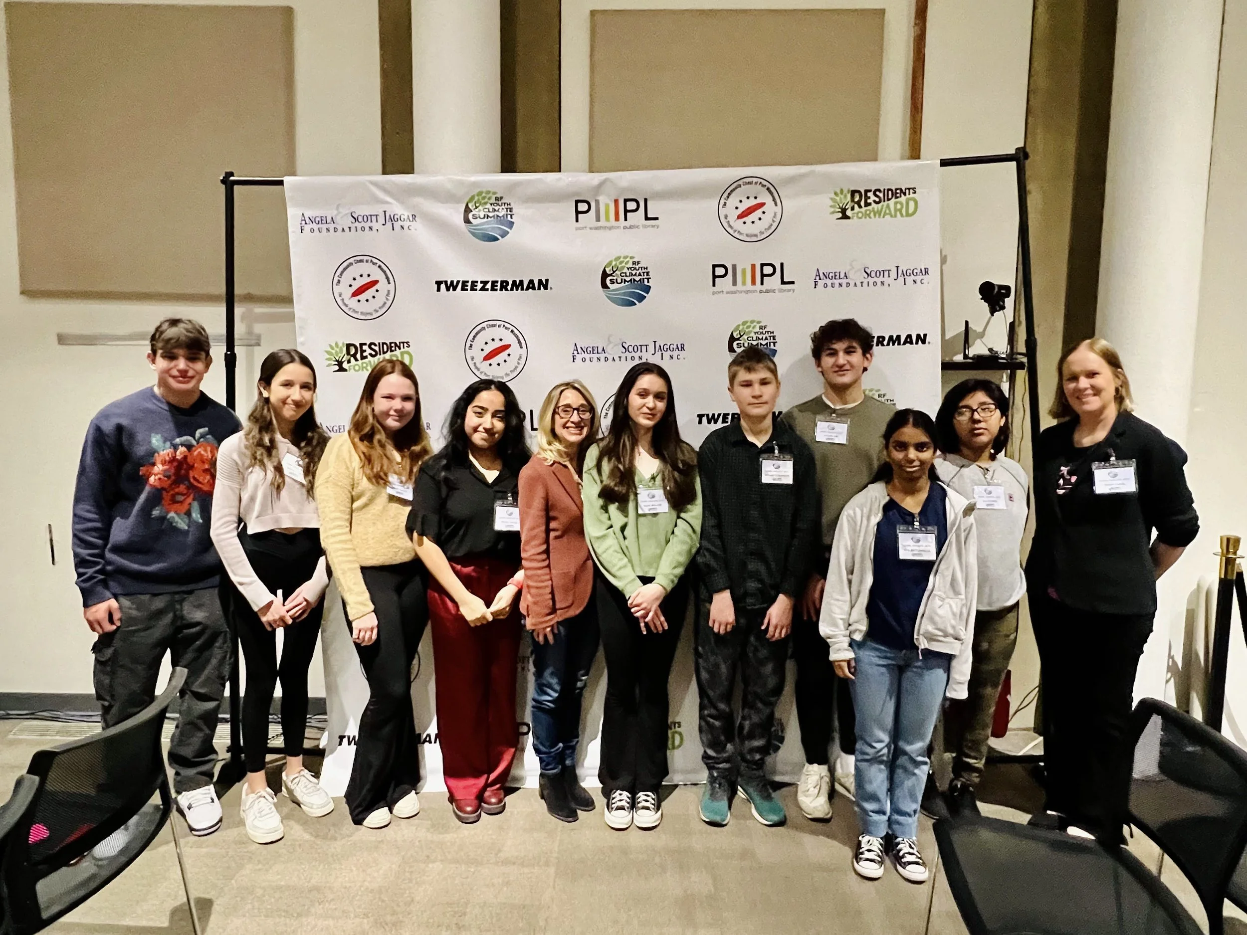 Group of twelve young people and an adult woman standing together in front of a backdrop with various logos at an indoor event.