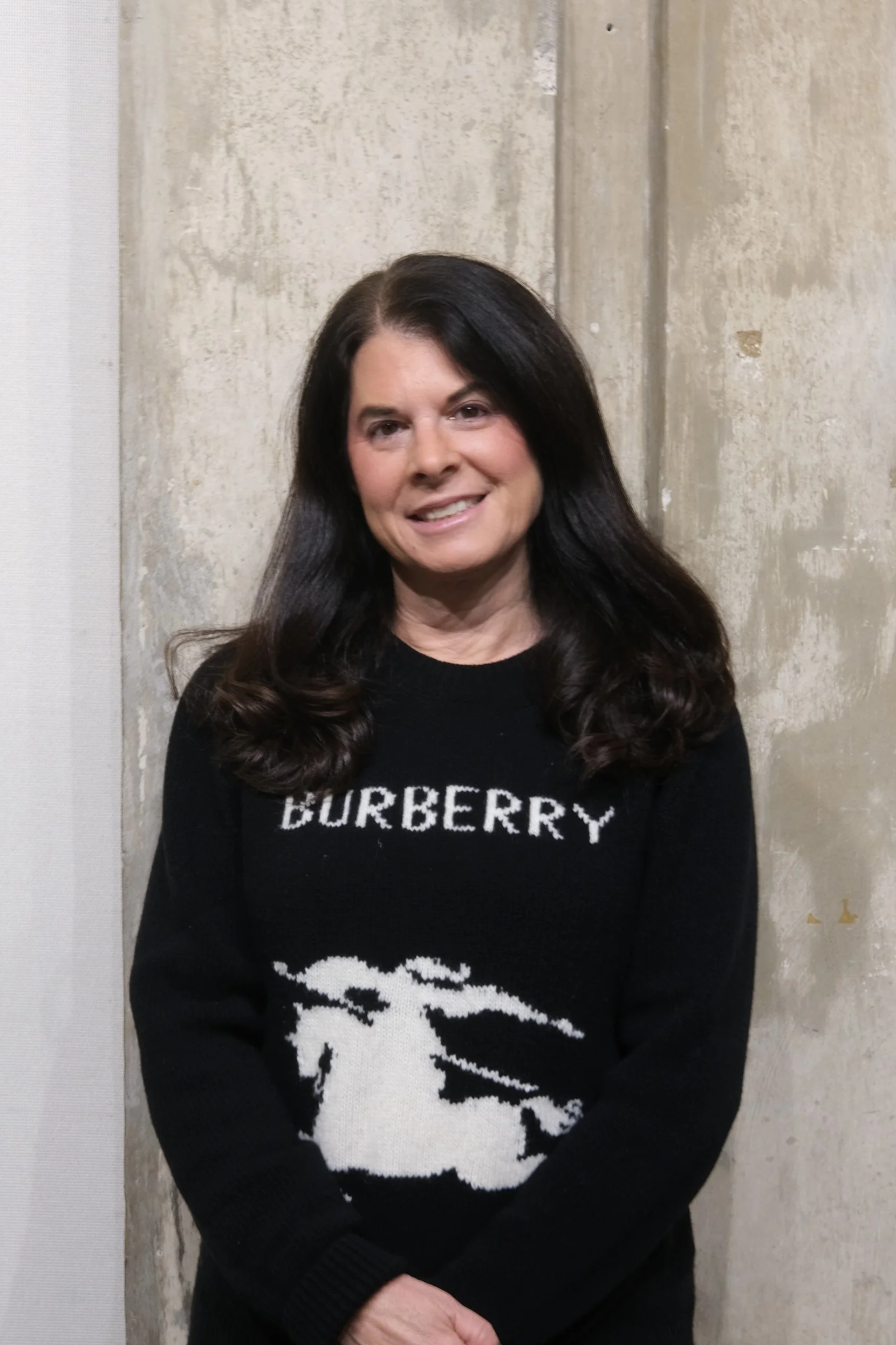 A woman with dark brown, wavy hair, wearing a black sweater with the word 'BURBERRY' and a white horse logo, smiling and standing against a textured beige wall.