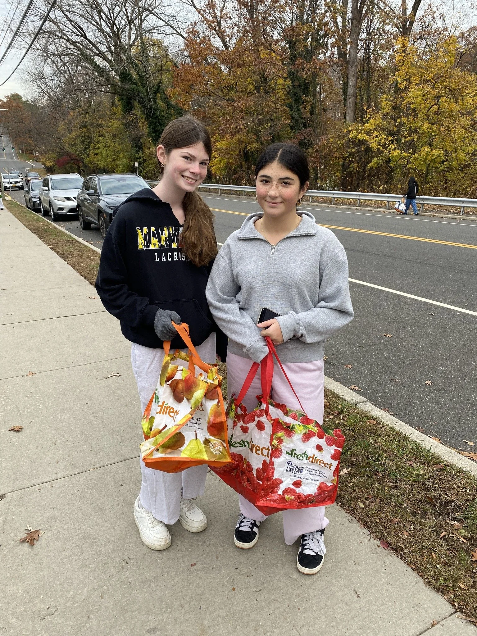 Two young women standing on a sidewalk holding grocery bags from FreshDirect, with parked cars and fall foliage in the background.