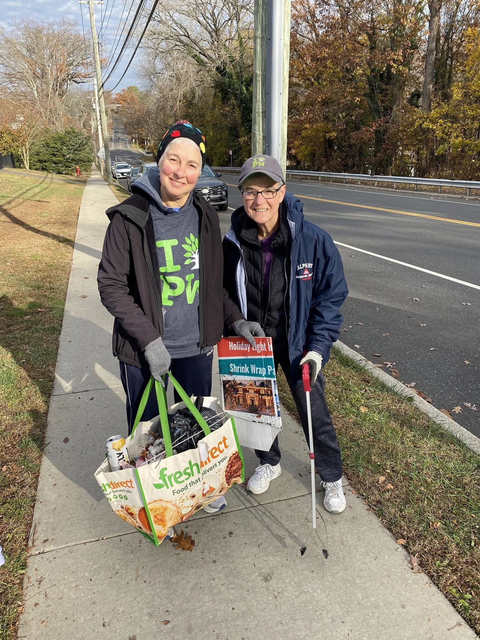 Two women standing on a sidewalk holding grocery bags and a flyer, smiling at the camera; one woman has a gray hat and glasses, and the other has a black beanie with colorful hearts; they are dressed warmly with jackets and gloves; a street and parke