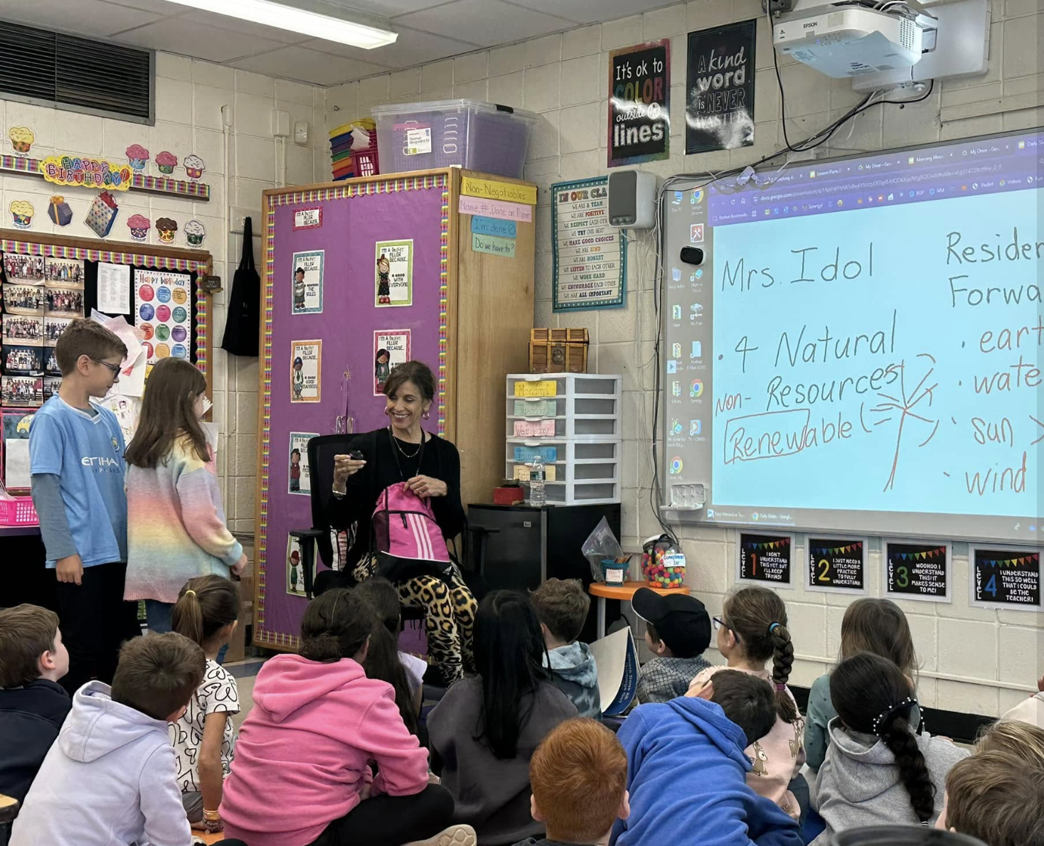 A classroom with students sitting on the floor and listening to a teacher who is standing and holding a pink backpack. The whiteboard displays notes about Mrs. Idol and renewable resources. There are various colorful posters and classroom decorations