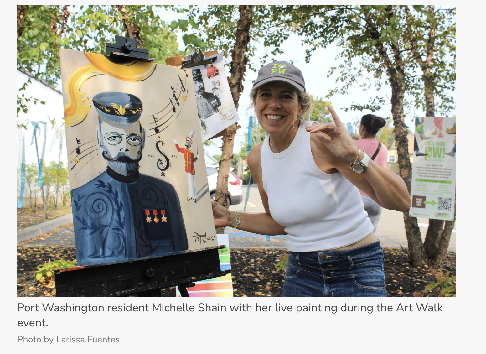Port Washington resident Michelle Shain smiling and waving next to her live painted portrait of a man with a beard and glasses, surrounded by musical notes, trees, and outdoor event signs during the Art Walk.