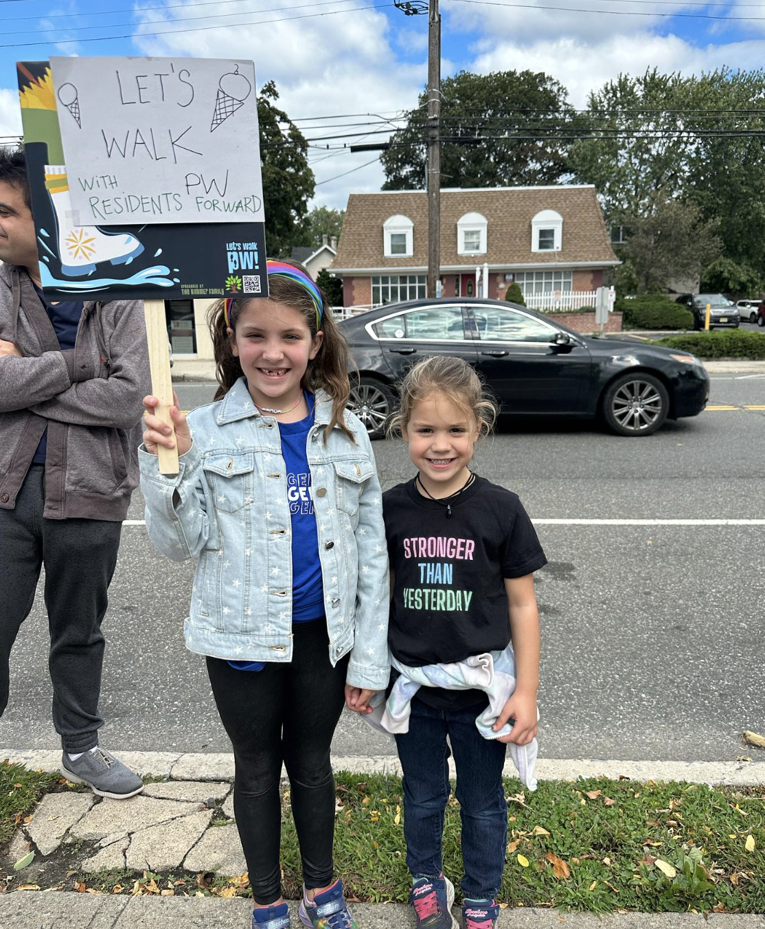 Two young girls standing on a sidewalk, holding a protest sign that reads 'Let's Walk with PW Residents Forward,' smiling at the camera, with houses and a car in the background.