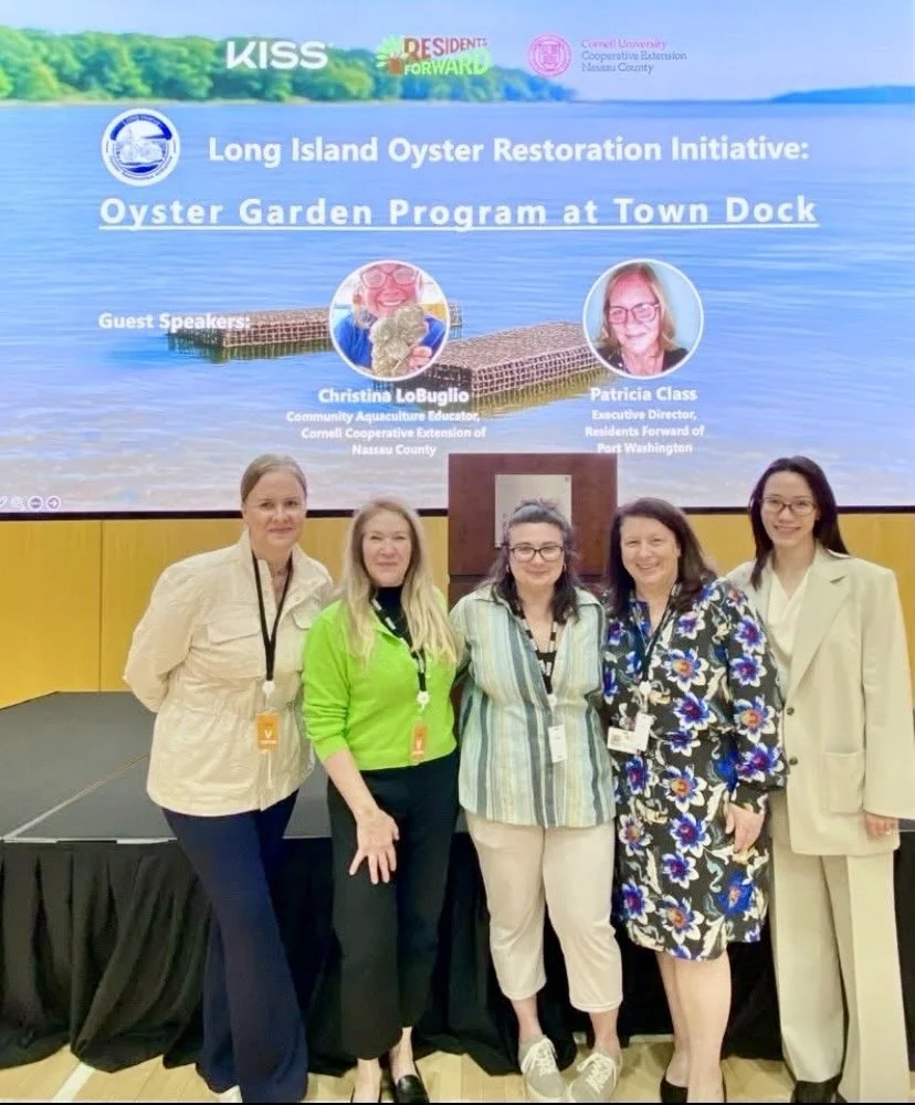 Group of five women standing in front of a presentation screen about the Long Island Oyster Restoration Initiative at Town Dock, with images of two guest speakers, Christina LoBuglio and Patricia Class.