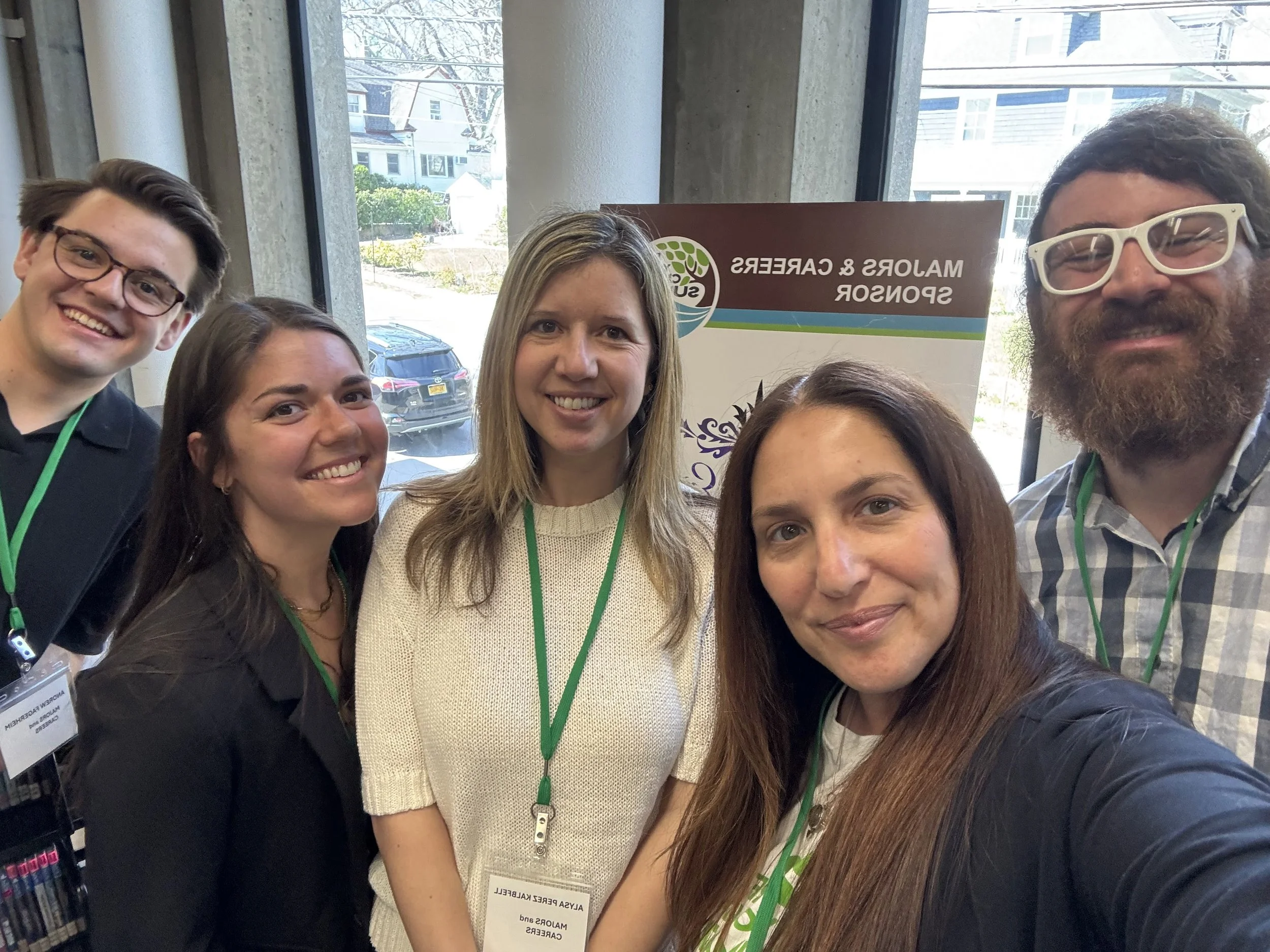 Group of five people taking a selfie at a conference, standing in front of a sign that reads, 'Malors & Carers Sponsored' with a blurred background of a building and parked cars outside.