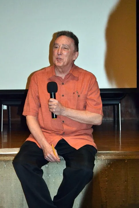 A man sitting on a stage holding a microphone and a yellow pencil, speaking during a presentation or talk.